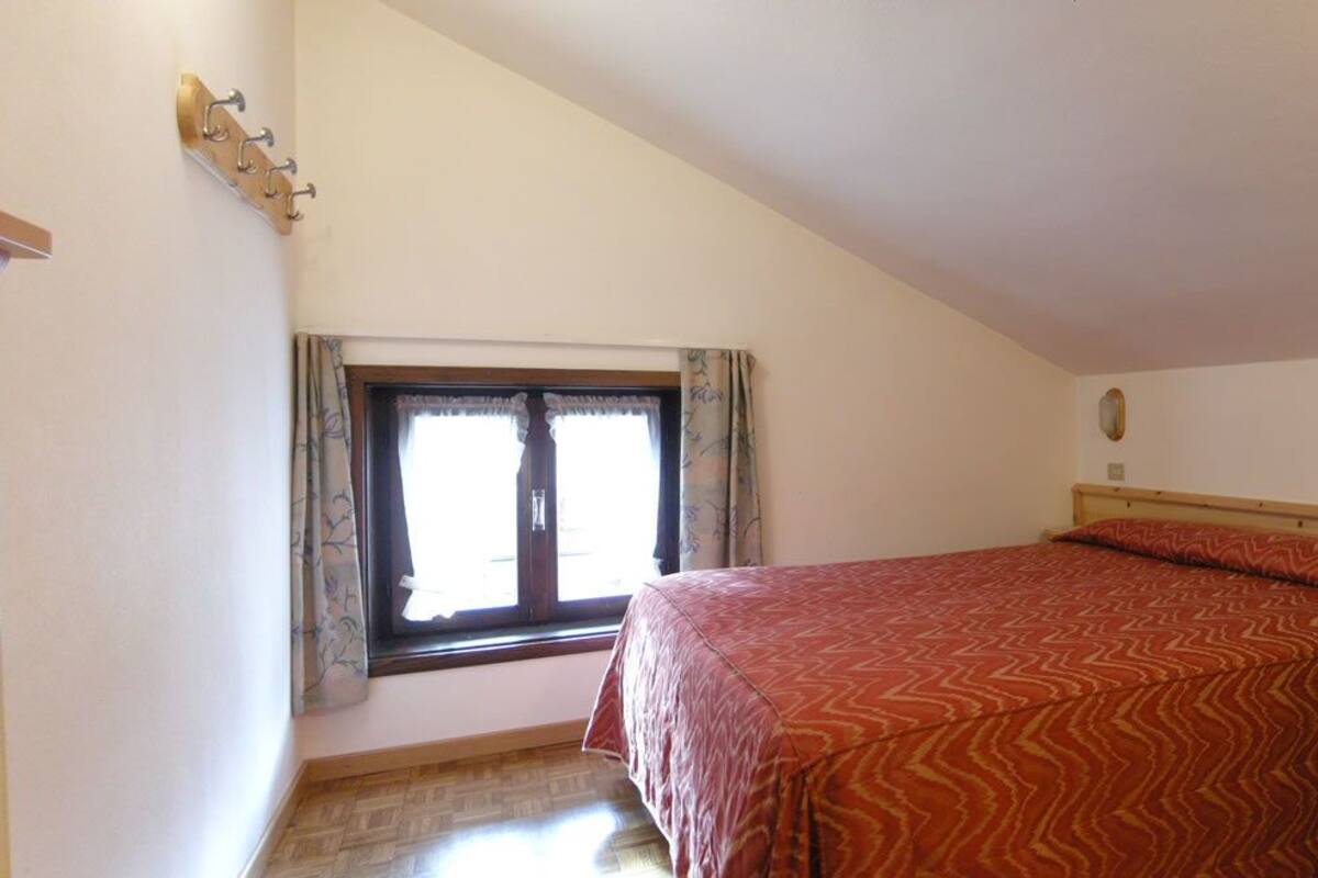 Ein Schlafzimmer mit einem Bett, einem Fenster mit Vorhängen, einem Holzschrank und einem Tisch mit einer Fernbedienung.