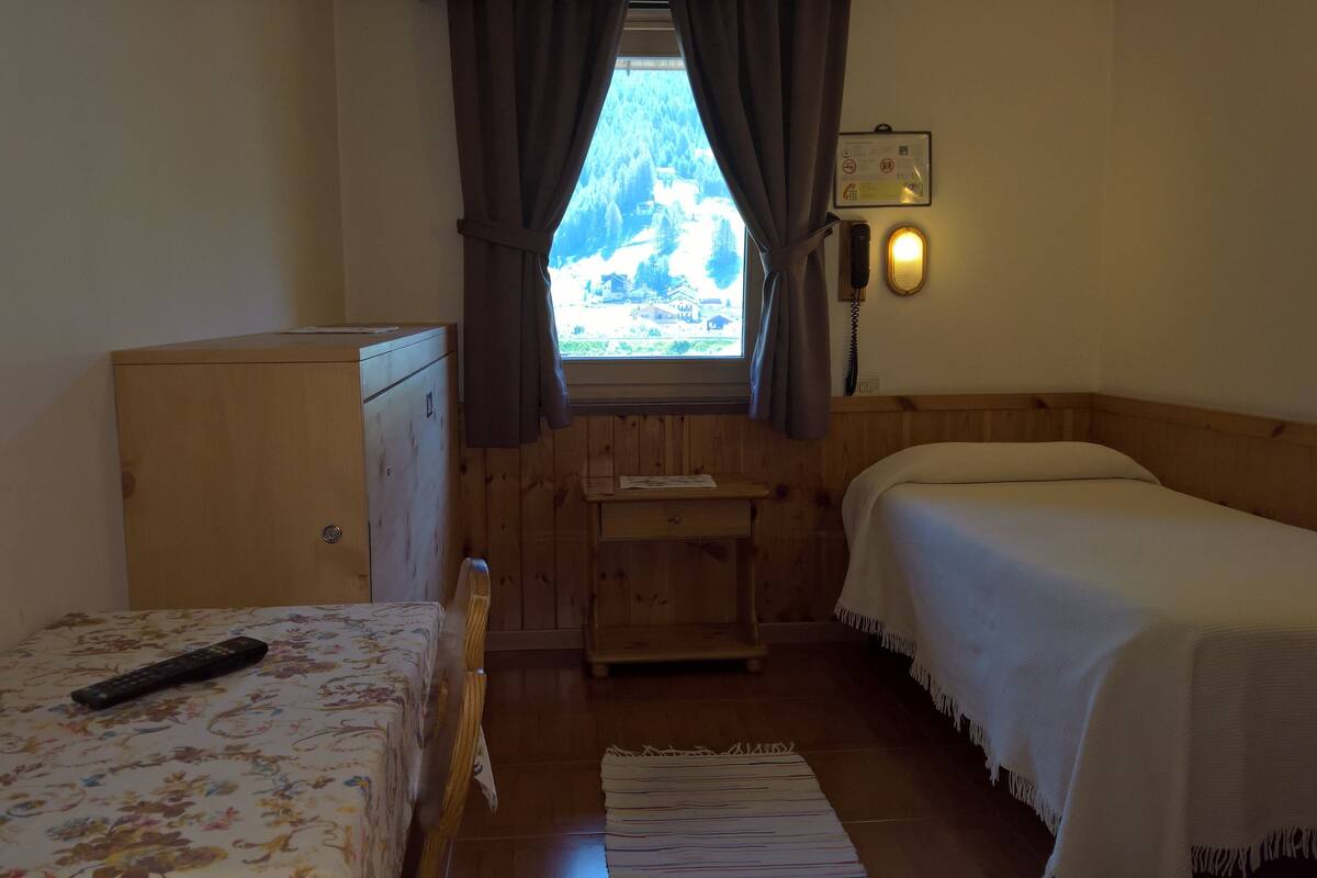 Ein kleiner, gemütlicher Raum mit einem Holzbett, einem Schreibtisch und einem Kleiderschrank. Ein Fenster mit Vorhängen, eine Wandlampe und eine Fernbedienung liegen auf dem Bett.