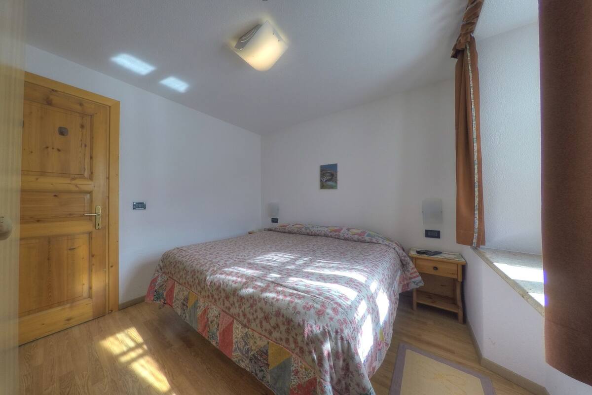 Ein Schlafzimmer mit Holztür, einem Bett mit floralem Überwurf, einem Nachttisch und einem Fenster mit Vorhängen.