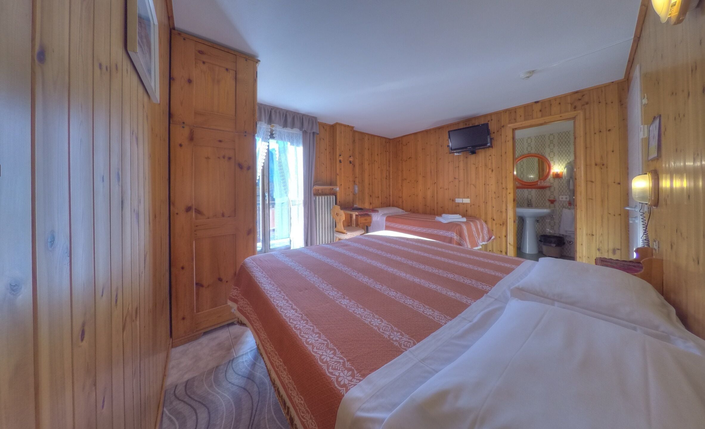Quarto de madeira com uma cama, uma escrivaninha, uma cadeira, uma televisão, um espelho e uma pia.