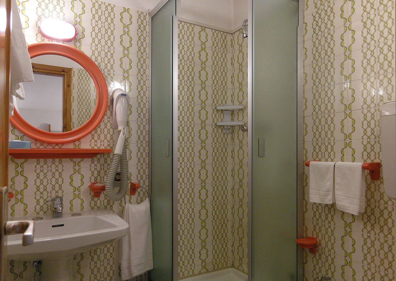 A bathroom with patterned wallpaper, a sink, a mirror, a hairdryer, and a shower.