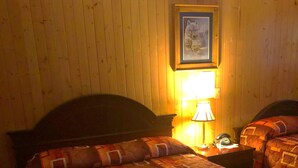 A room with a wooden paneled wall, a bed with a patterned bedspread, a nightstand with a lamp, and a framed picture on the wall. - Alta Vista Motel (Baldwin)