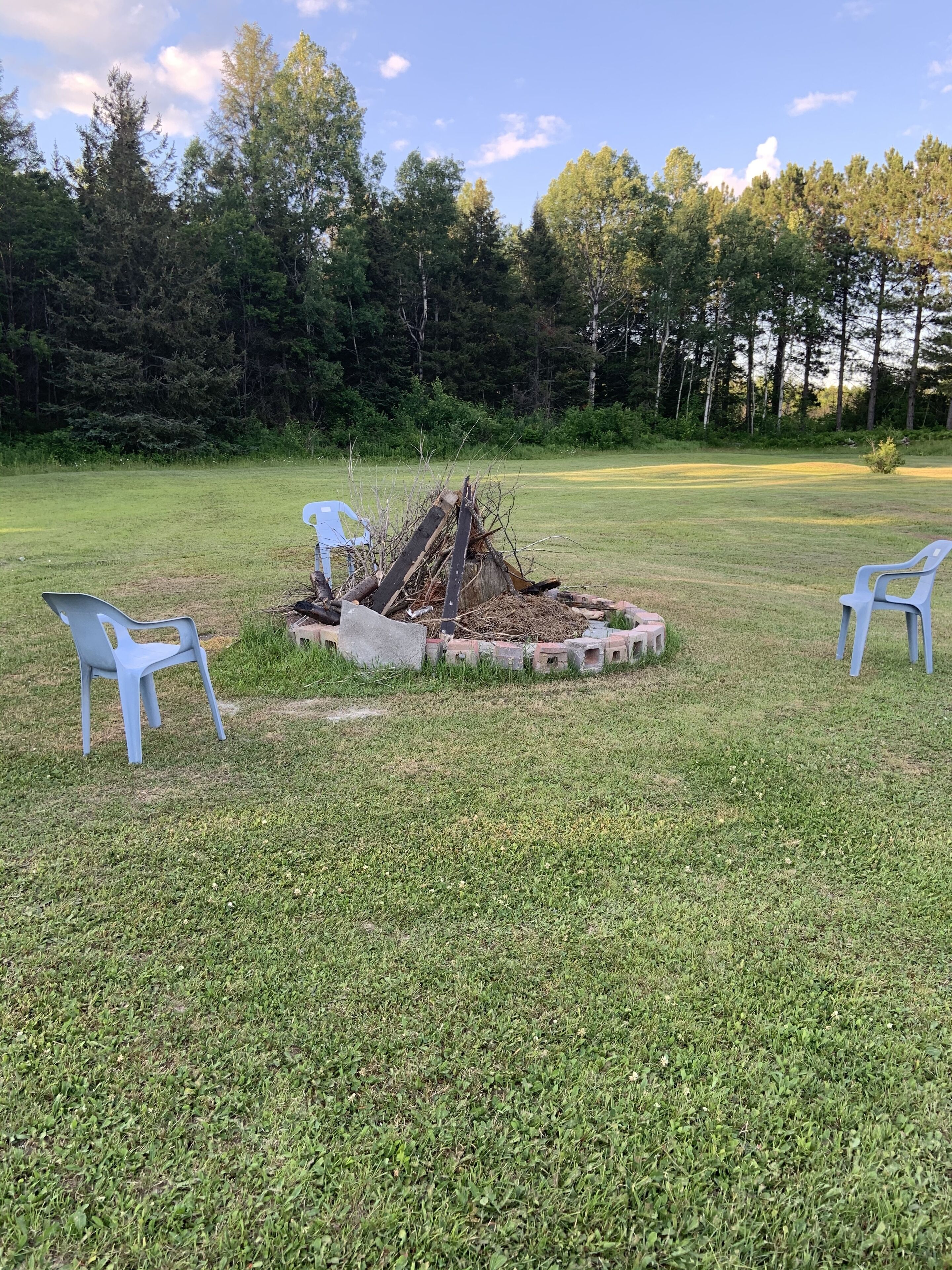 A grassy outdoor area with a fire pit surrounded by concrete blocks, and two white plastic chairs.