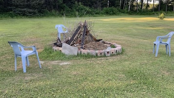 A grassy outdoor area with a fire pit surrounded by concrete blocks, and two white plastic chairs.