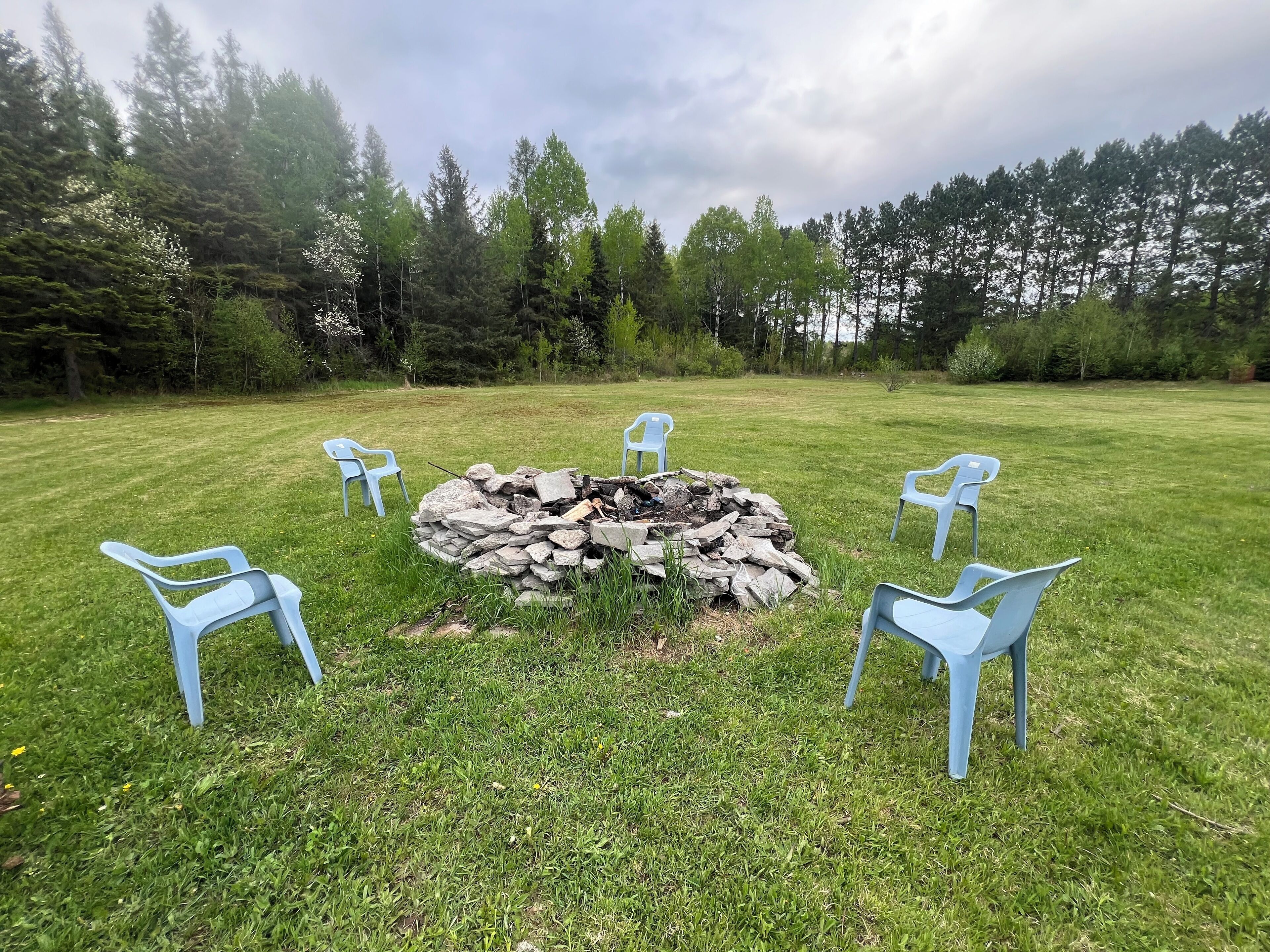 A stone fire pit surrounded by four blue plastic chairs in a grassy outdoor area with trees in the background.
