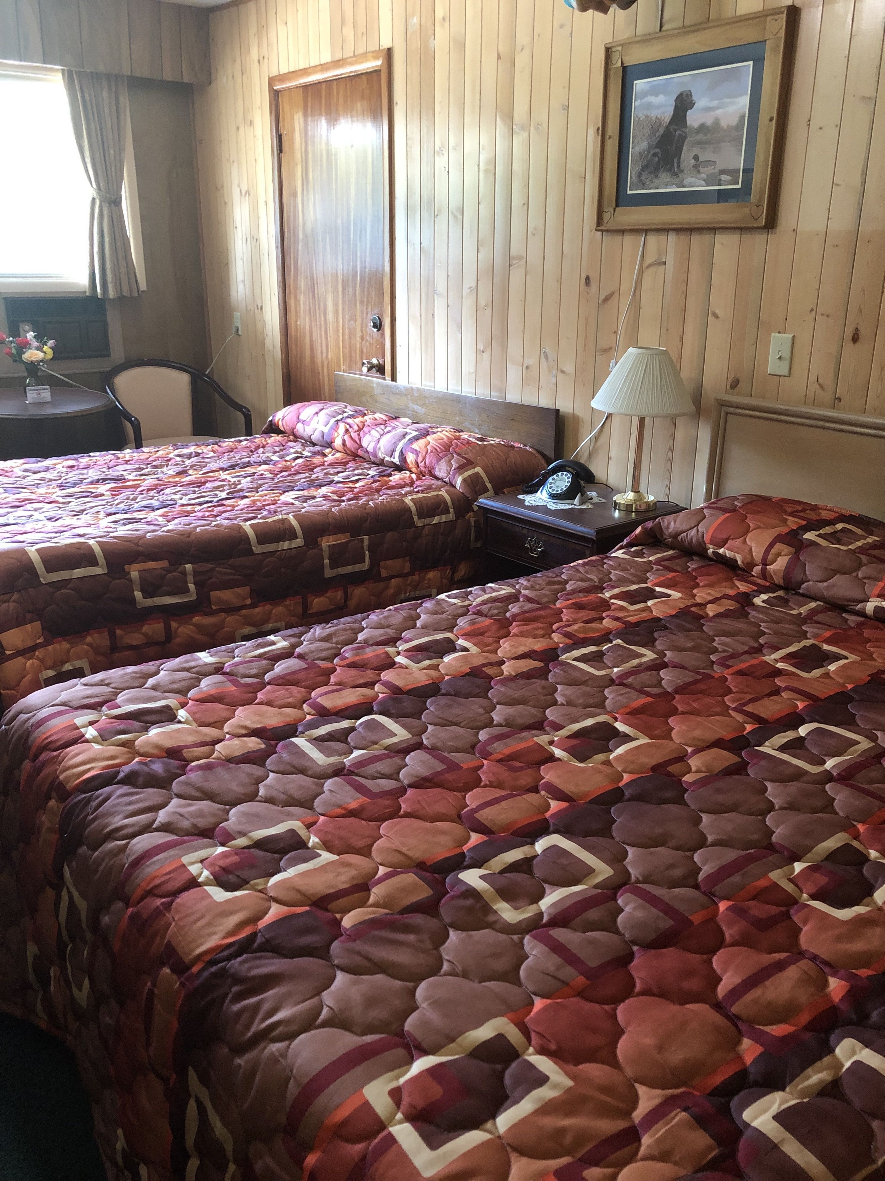 Una habitación con dos camas, paredes de madera, un cuadro enmarco, un escritorio con lámpara y una silla.