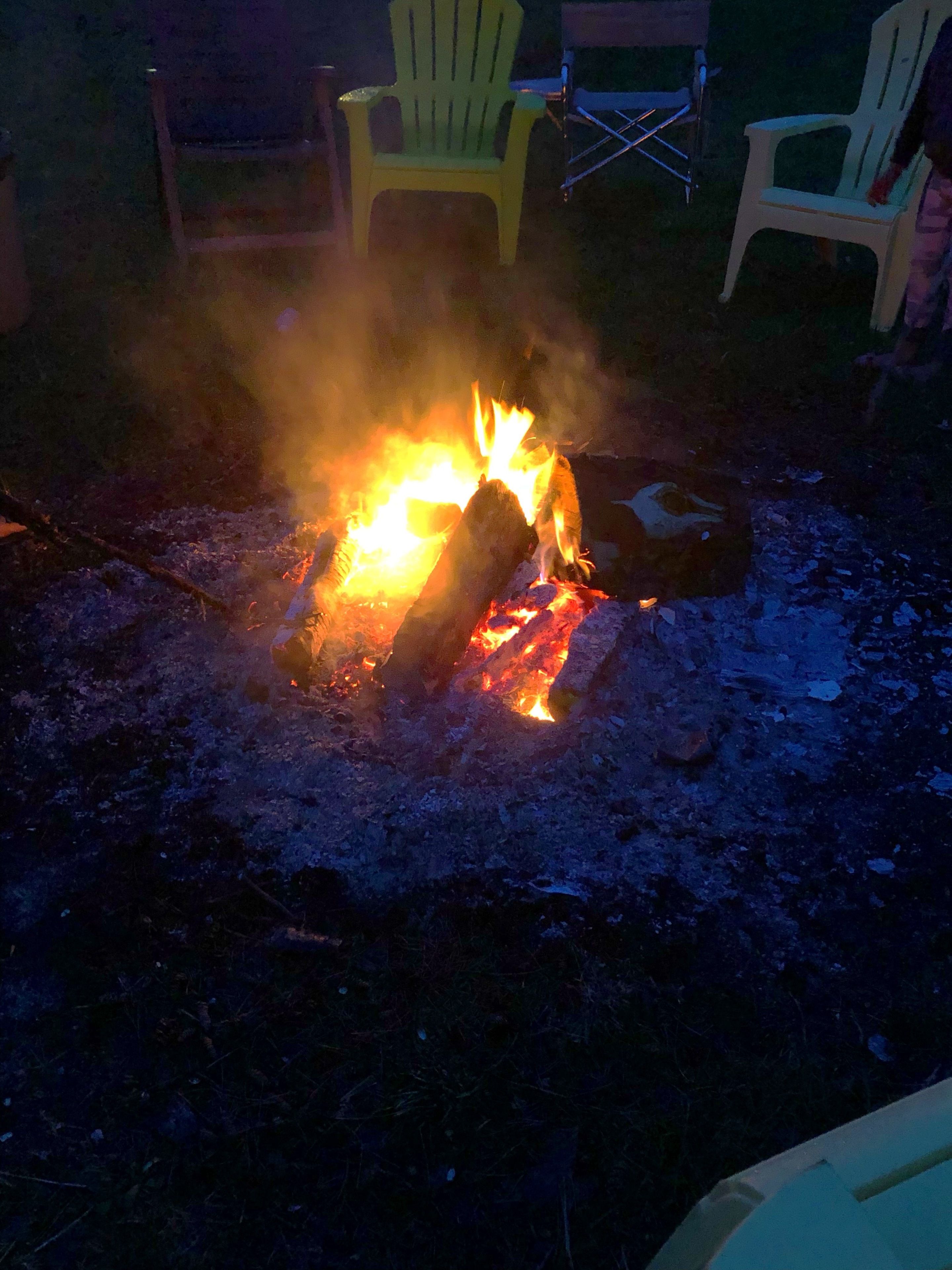 Un foc de campament amb llamenes i fum visibles, envoltat de cadirees i una nevera.