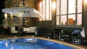 Une piscine entourée de chaises longues et de parasols, le tout bordé d’un bâtiment doté de grandes fenêtres et d’un balcon.
