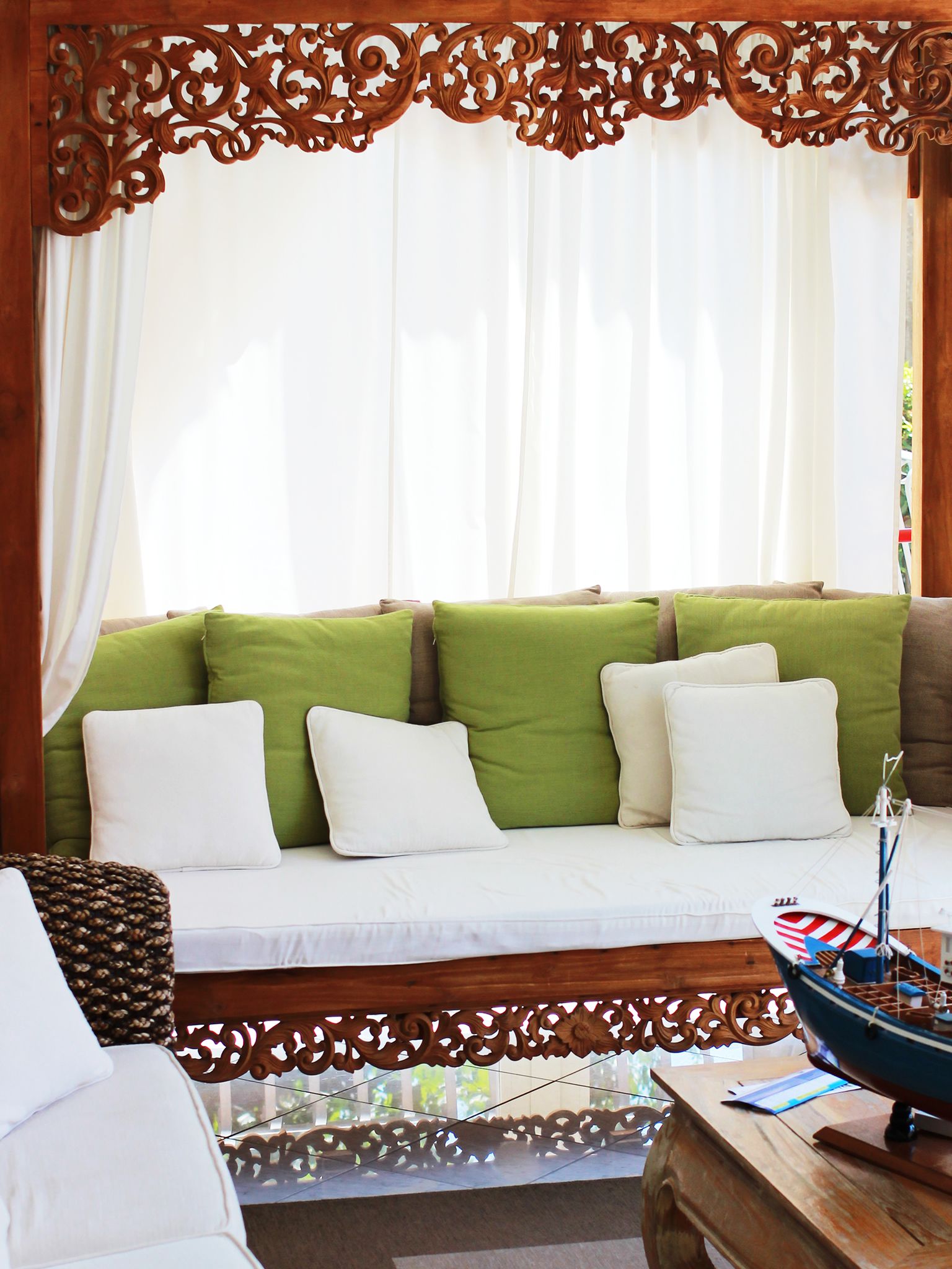 A wooden-framed window with white curtains, a sofa with green and white cushions, and a small table with a model ship.