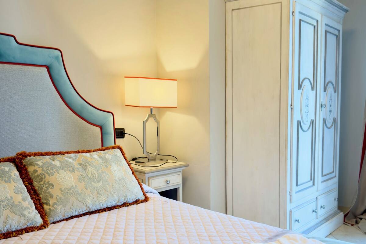 A neatly made bed with a quilted headboard, a bedside table with a lamp, and a white wardrobe.