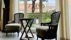 Une pièce avec deux fauteuils en osier, une petite table et une vue sur une piscine et des bâtiments, le tout visible à travers de grandes fenêtres.