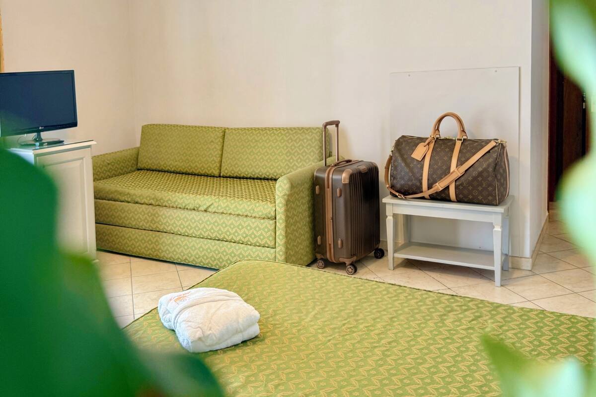 A room with a green patterned sofa, a white bedside table with a Louis Vuitton bag, a suitcase, and a television on a stand.