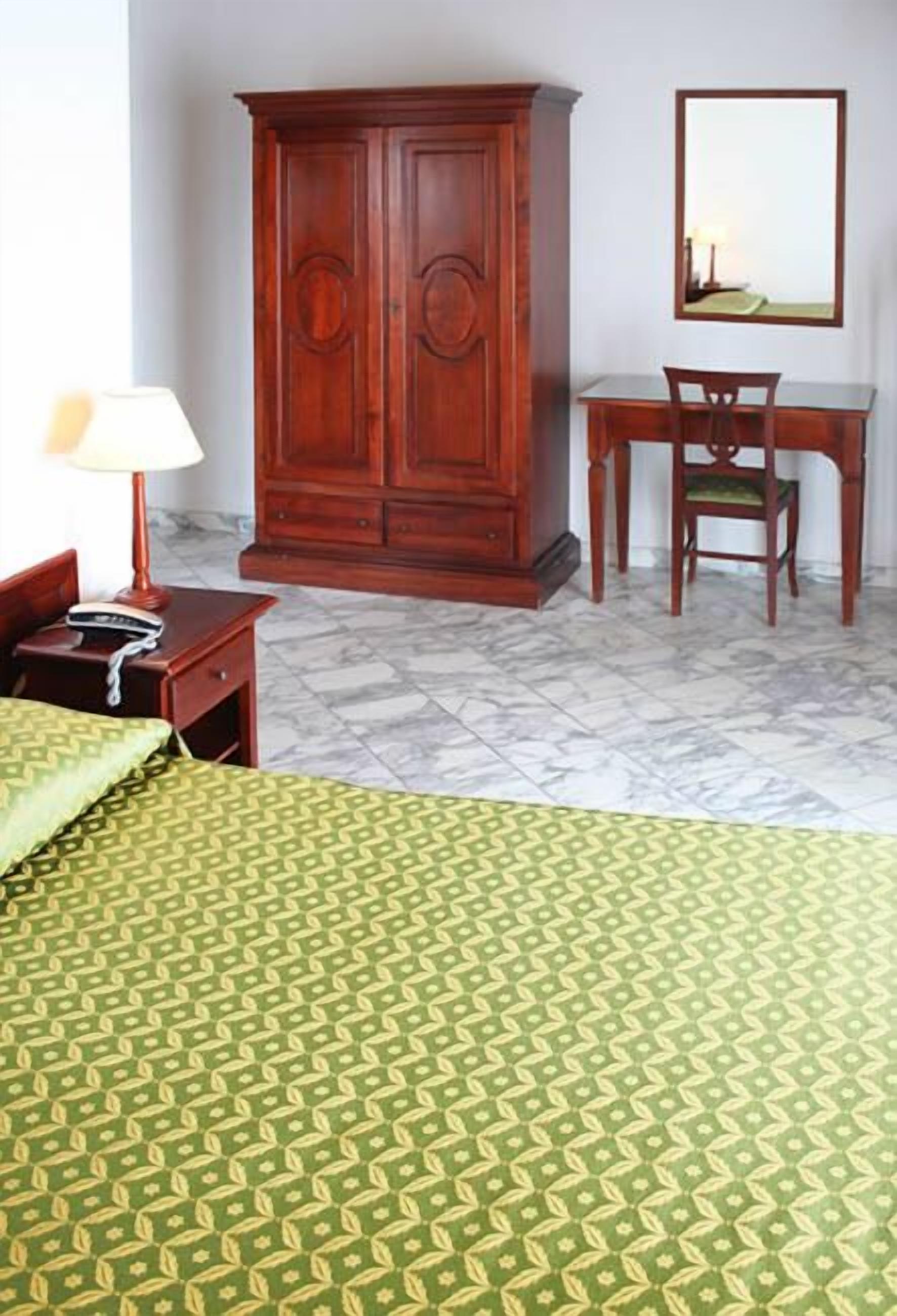 A hotel room with a bed, a wooden wardrobe, a desk with a chair, and a mirror on the wall.