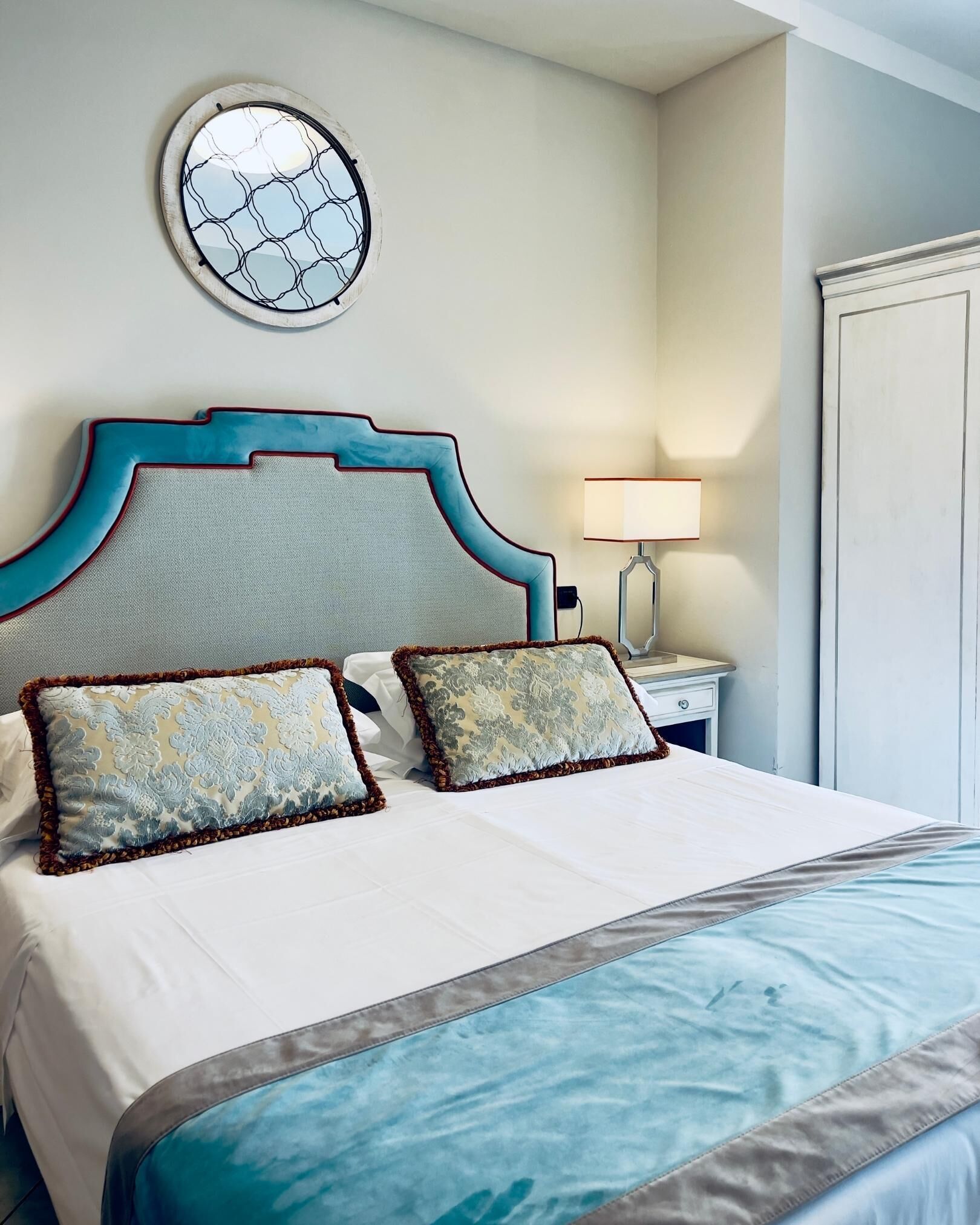 A neatly made bed with a blue and white bedspread, two decorative pillows, a round wall mirror, and a bedside lamp.