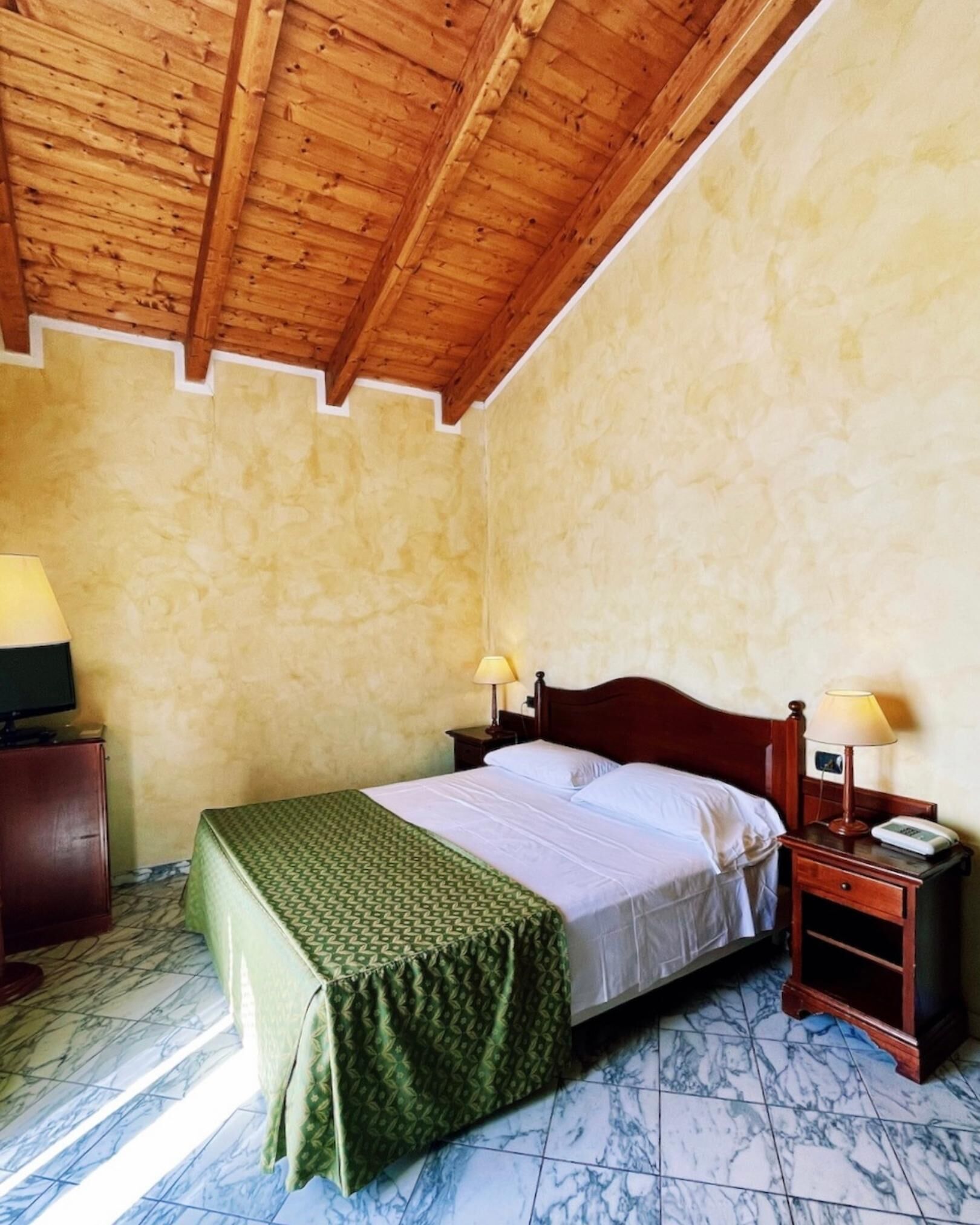 A single bed with a green patterned bedspread, two bedside lamps, a wooden nightstand, and a television on a wooden cabinet.