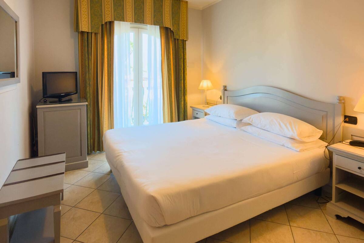 A hotel room with a bed, bedside tables, a TV, curtains, and a ceiling light.