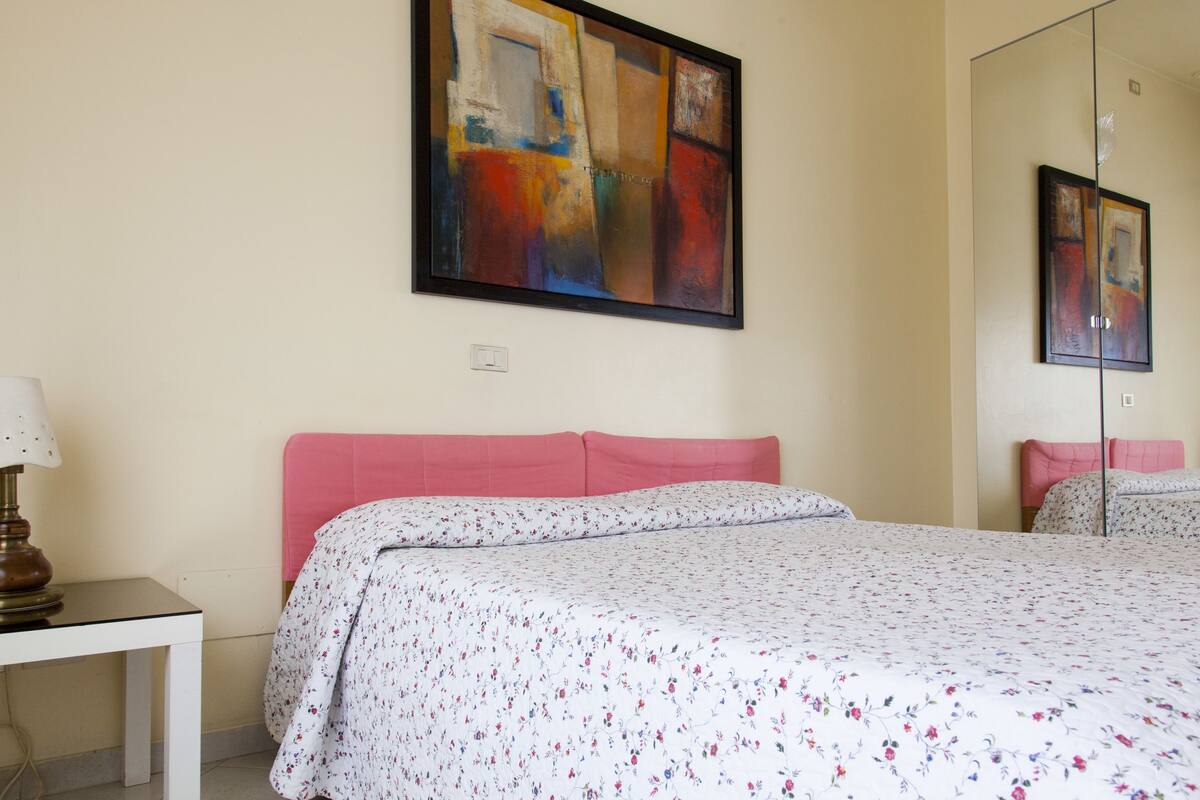 A hotel room with a bed, a bedside table, a lamp, and a large abstract painting on the wall.