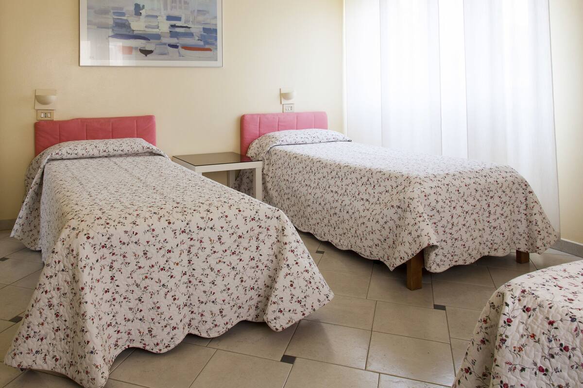 A hotel room with two single beds, a small table, and a wall-mounted light fixture.