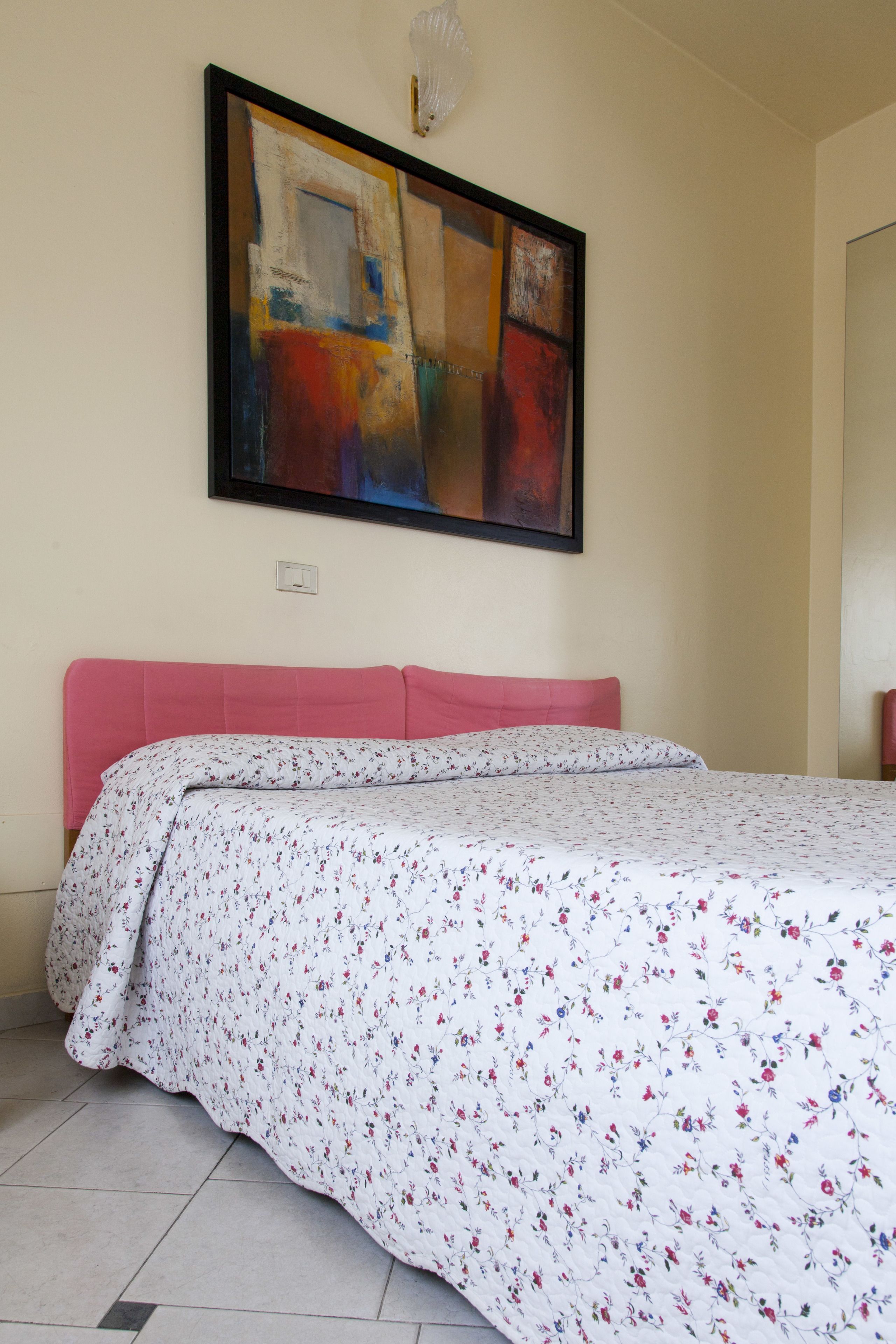 Een slaapkamer met een bed, een roze hoofdbord, een bloemenprint deken en een groot abstract schilderij aan de muur.