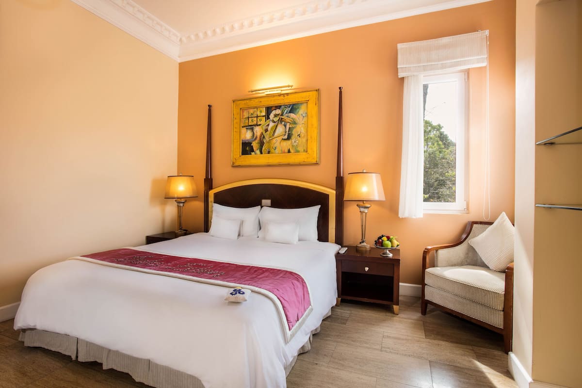 A hotel room with a large bed, two bedside tables, a chair, a window with curtains, and a painting on the wall.