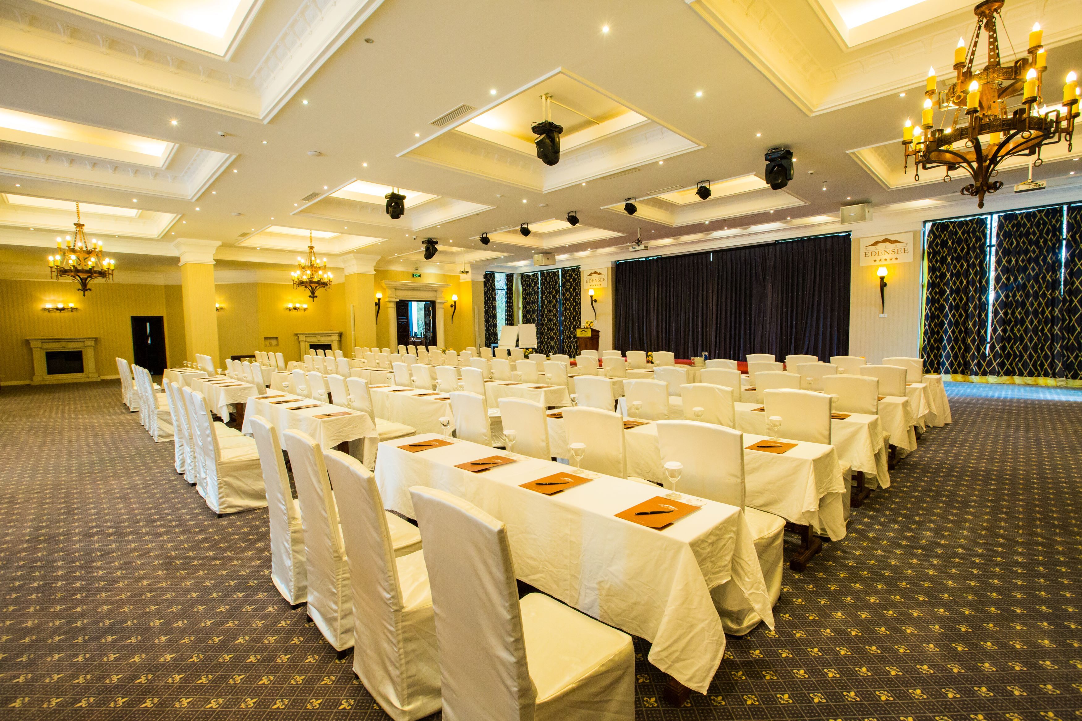A conference room with rows of white chairs, tables with name cards, a stage with a curtain, and a chandelier.