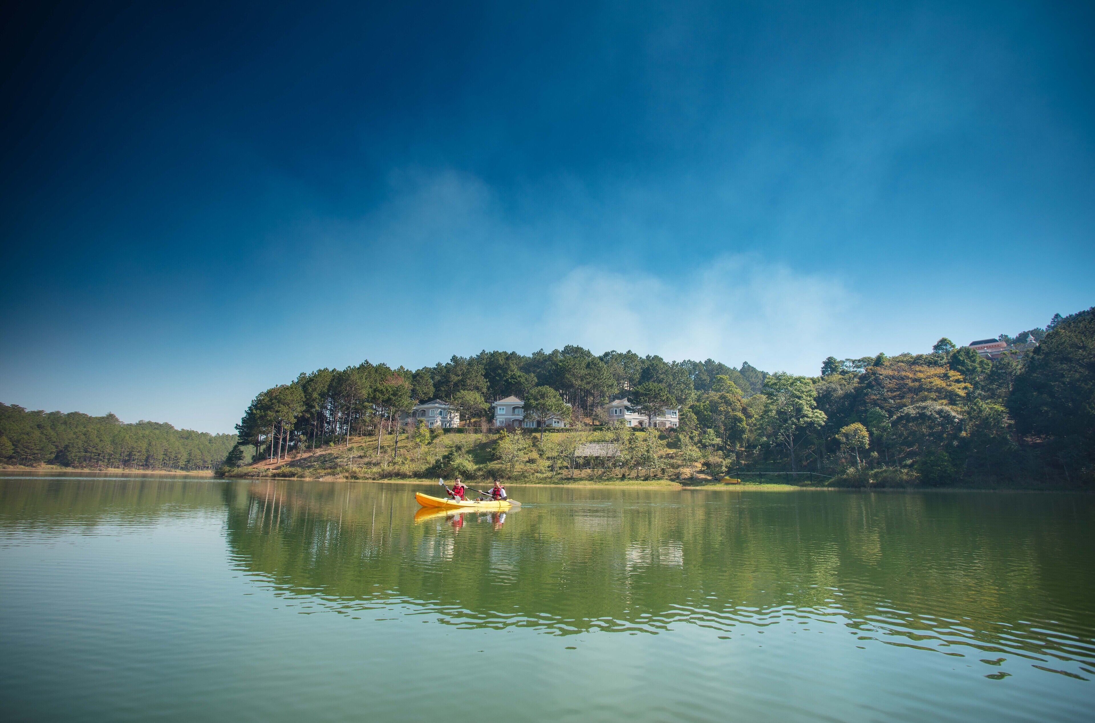 Um lago tranquilo com um caiaque e duas pessoas remando. Casas são visíveis na margem.