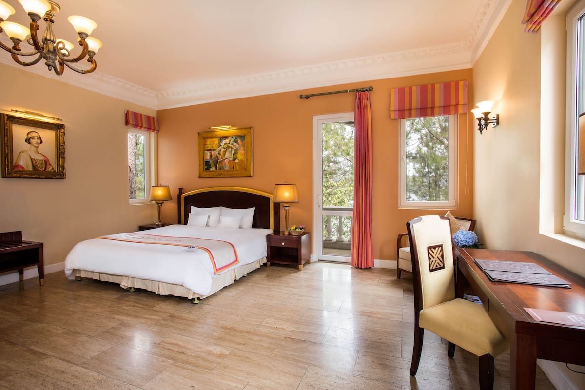 A hotel room with a large bed, two bedside tables with lamps, a desk with a chair, and a chandelier.