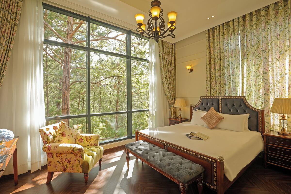 A spacious bedroom with a large window, a grand bed, a bench, and a chair.