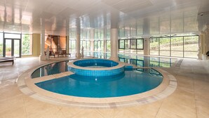 Una amplia piscina interior con un jacuzzi, rodeada de grandes ventanales y suelo de piedra.