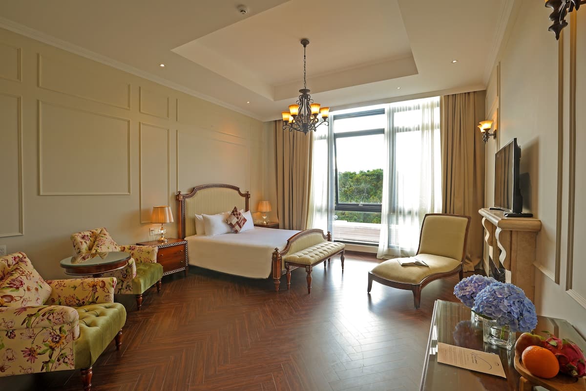 A spacious hotel room with a large bed, a sitting area with a sofa and armchairs, a dining table with a vase of flowers, and a view of greenery through the window.