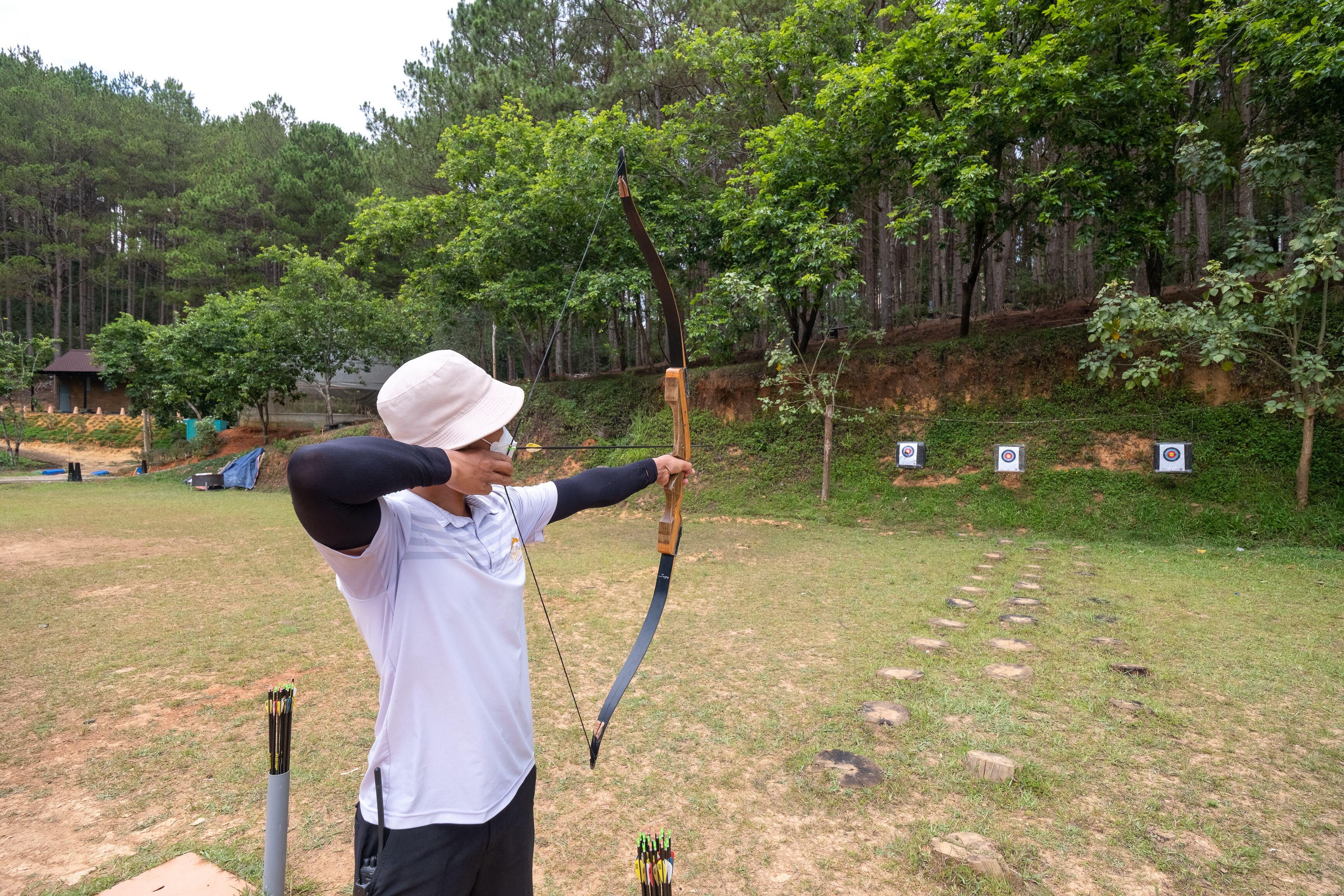 An outdoor archery range with targets and a person in a white shirt and hat aiming a bow.