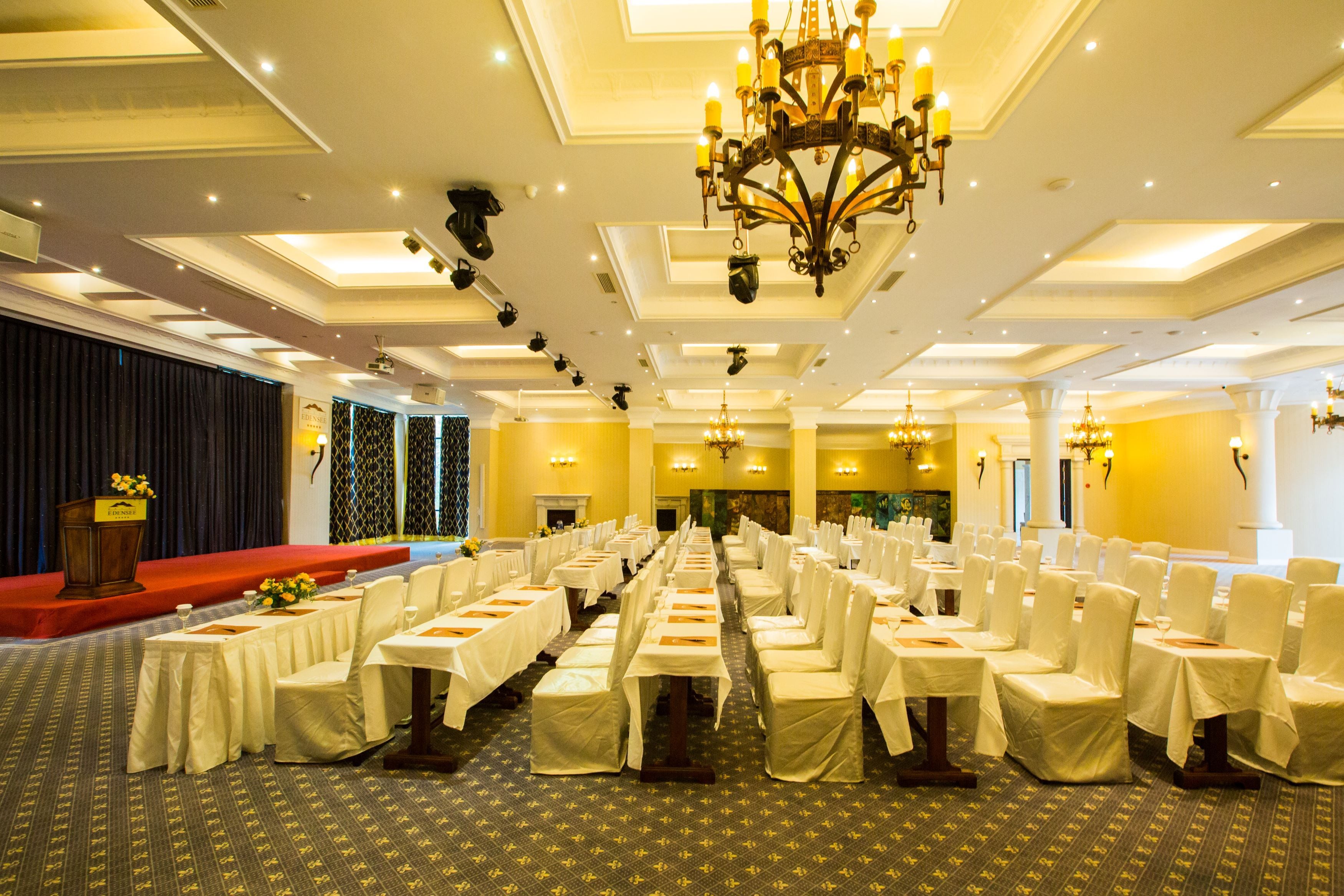 A banquet hall with rows of tables set for dining, a stage with a podium, and a chandelier.