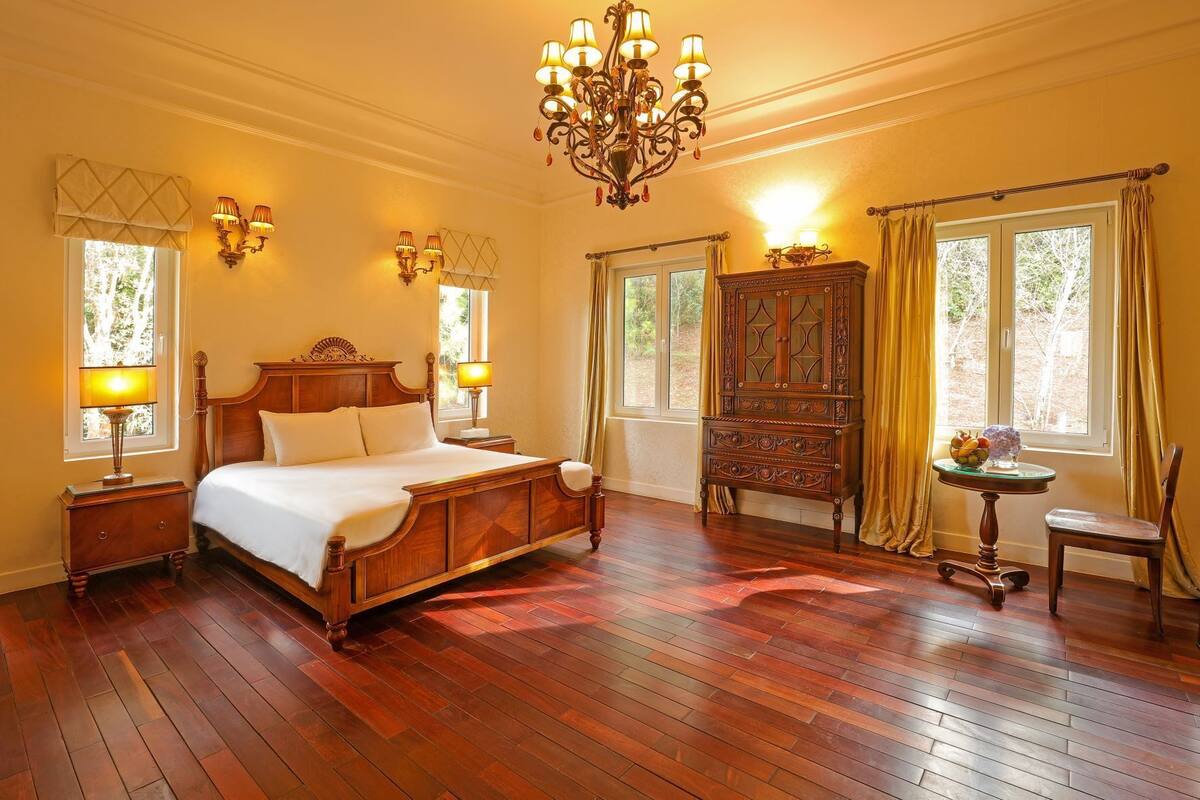 A spacious bedroom with a large wooden bed, two bedside lamps, a chandelier, a wooden cabinet, and a small table with a vase.