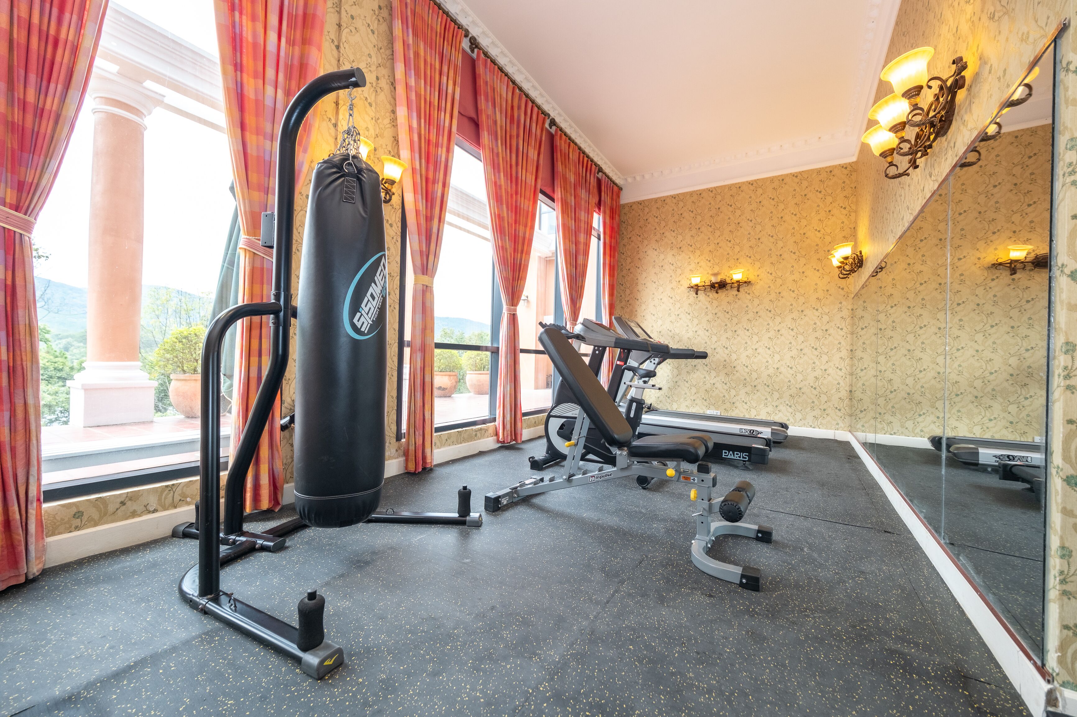 Ein Fitnessraum mit Trainingsgeräten, großen Fenstern mit Vorhängen und einem Spiegel, der den Raum widerspiegelt.