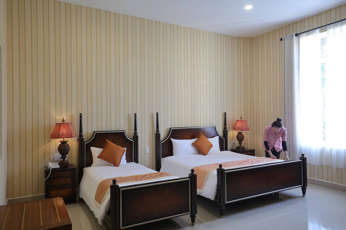 A hotel room with two beds, a nightstand with a lamp, and a wooden bench.