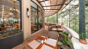 An outdoor dining area with wicker furniture, potted plants, and a view of the forest. - Dalat Edensee Lake Resort & Spa (Xuan Huong - Da Lat)