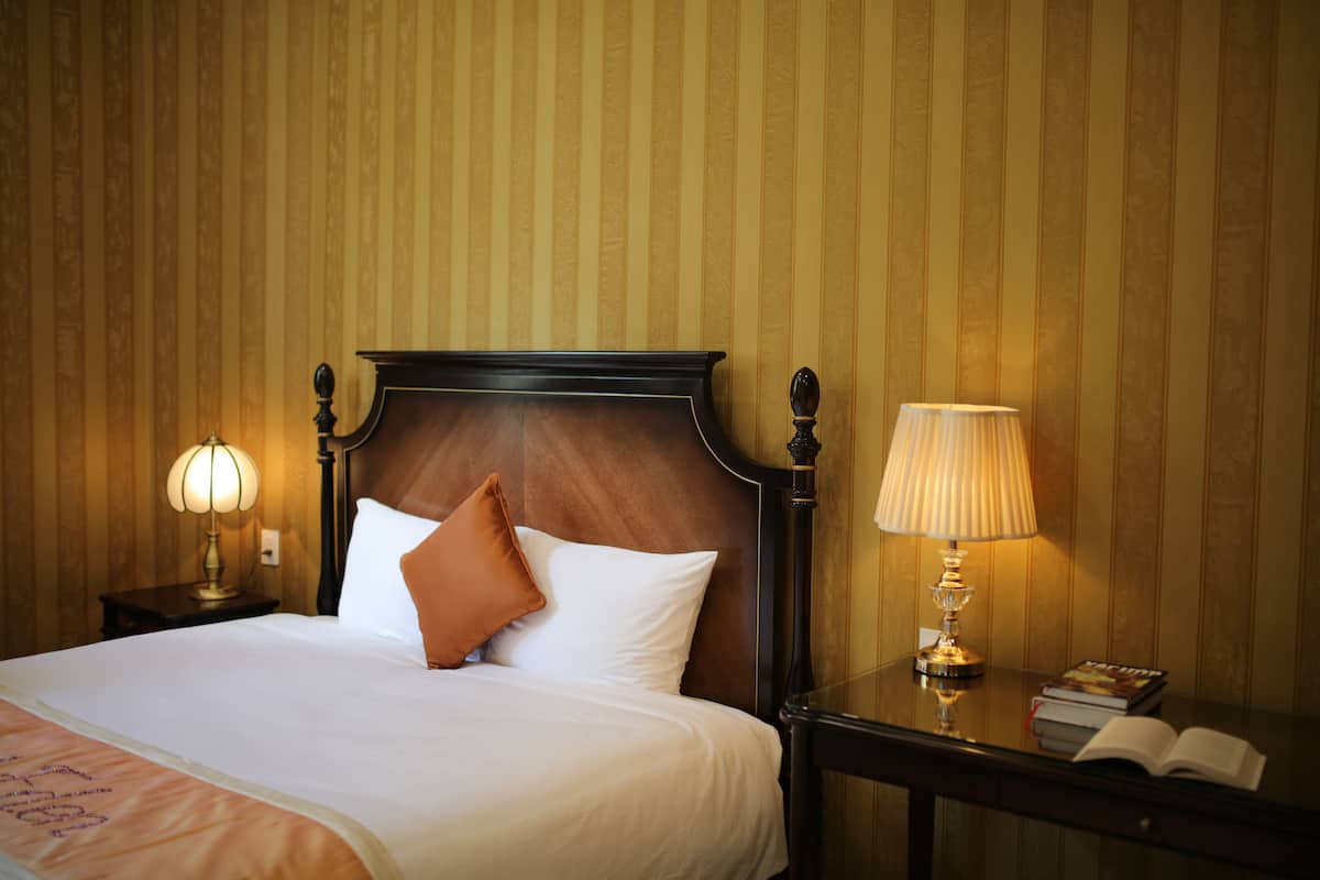 A neatly made bed with white linens and a brown pillow, flanked by two bedside tables with lamps and books.