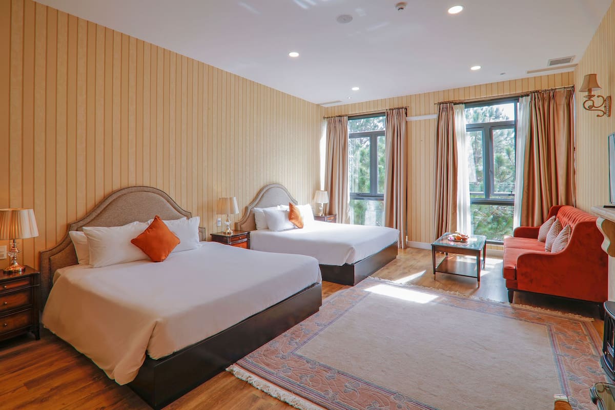 A hotel room with two beds, a red sofa, a small table, and a large window with curtains.