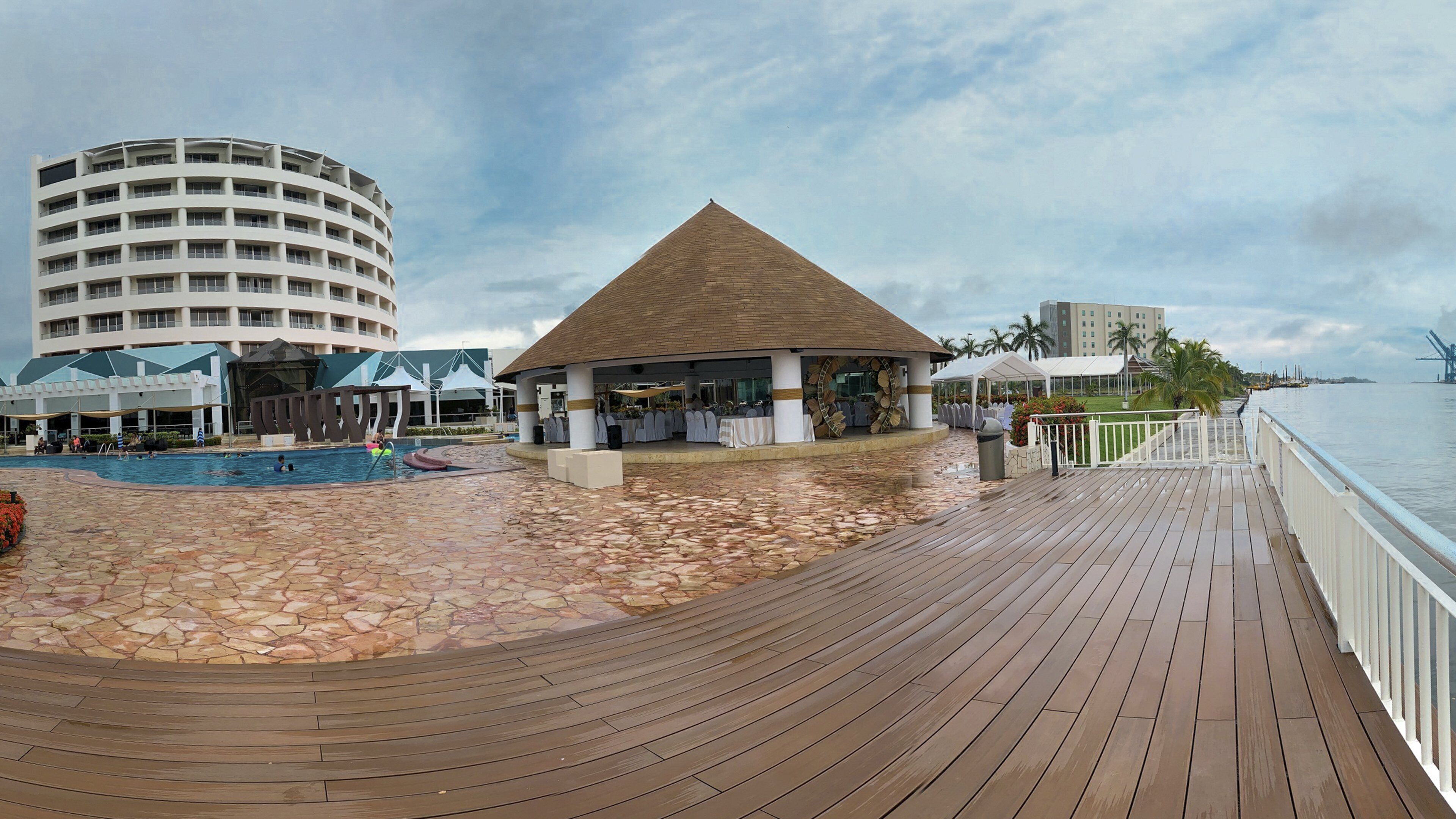 Un resort con piscina, zona de comedor cubierta y terraza de madera con vista al agua.