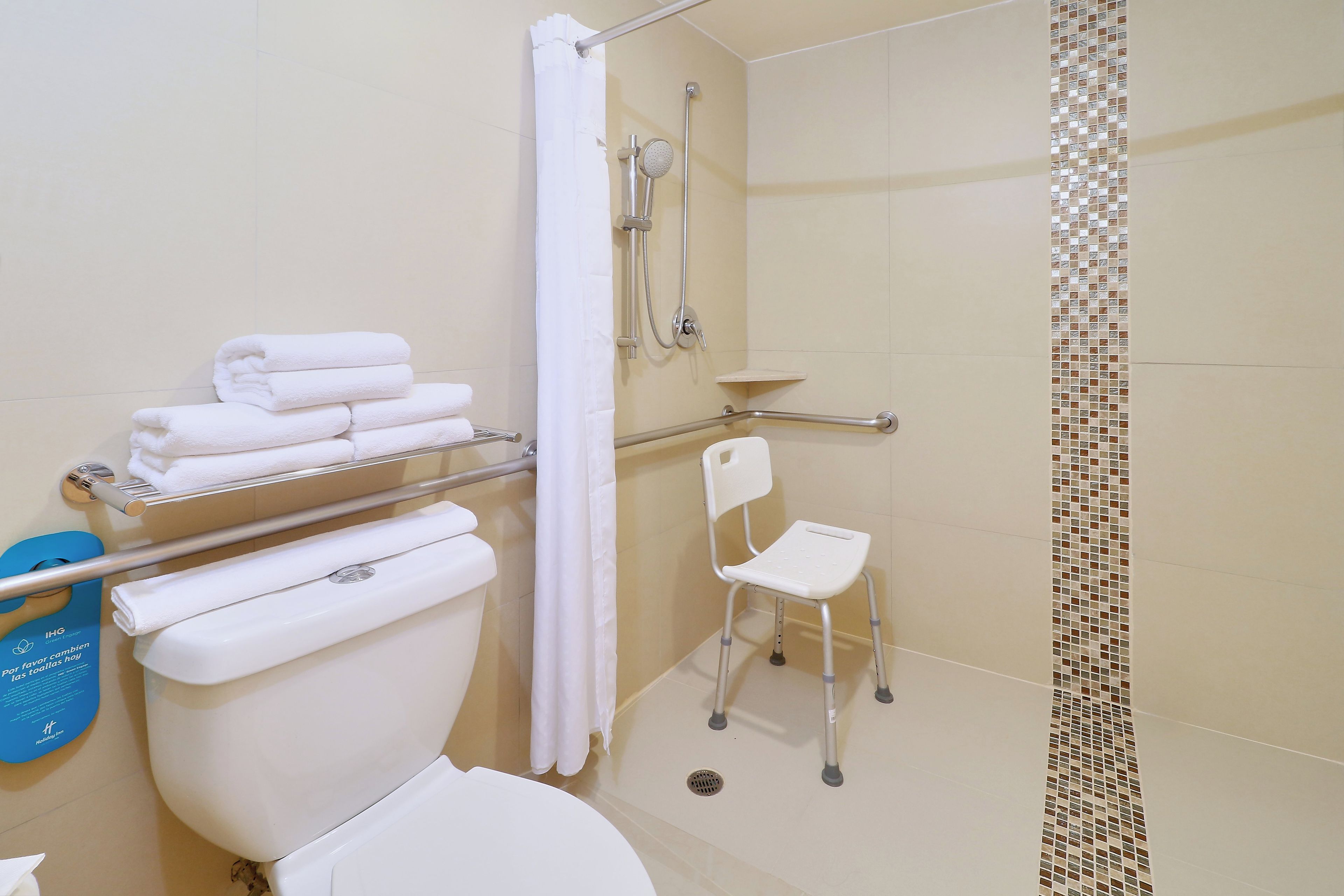 A bathroom with a toilet, a shower with a grab bar, a white chair, and a stack of white towels.