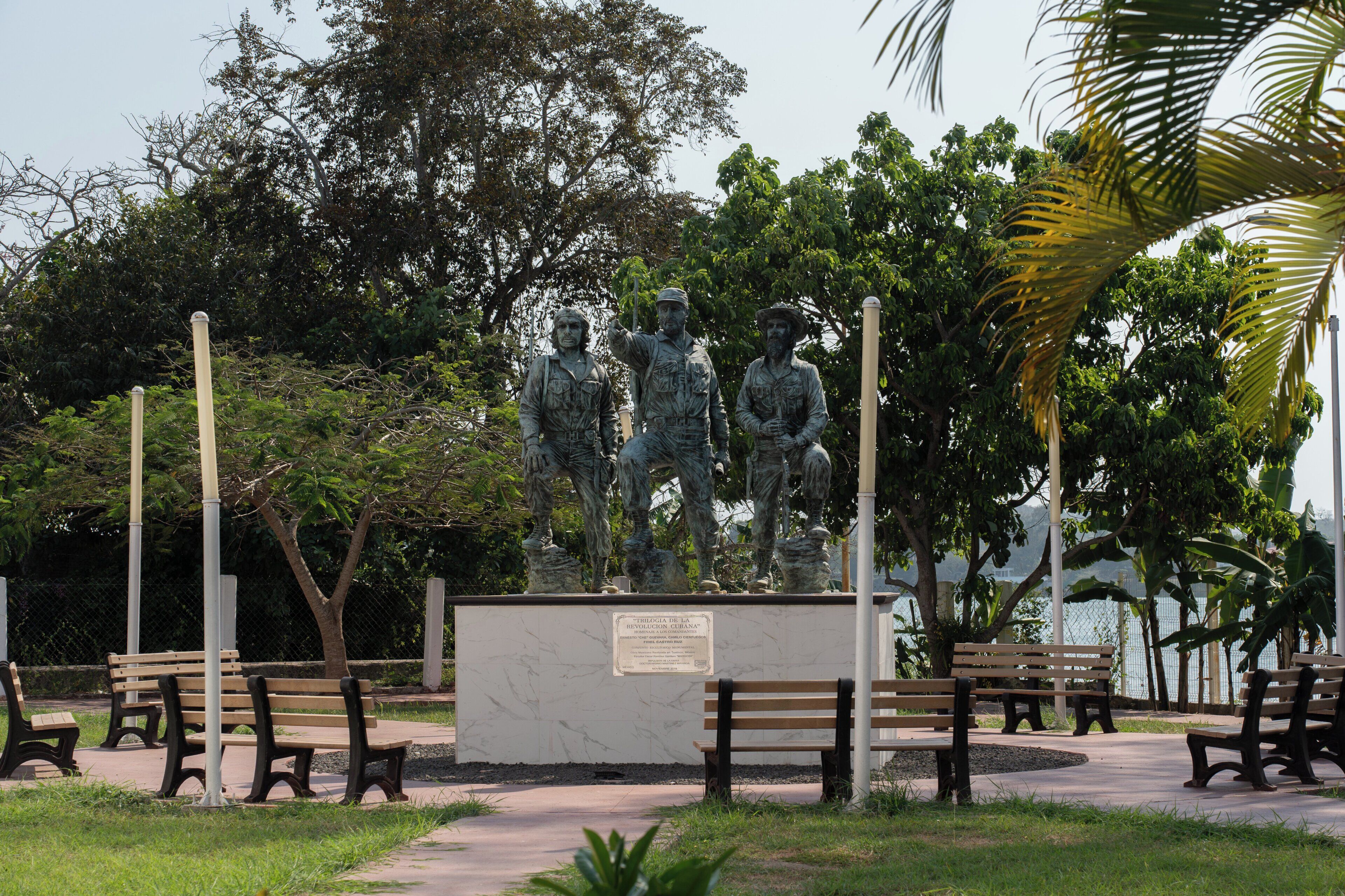 A statue of three figures in a park with benches and trees.