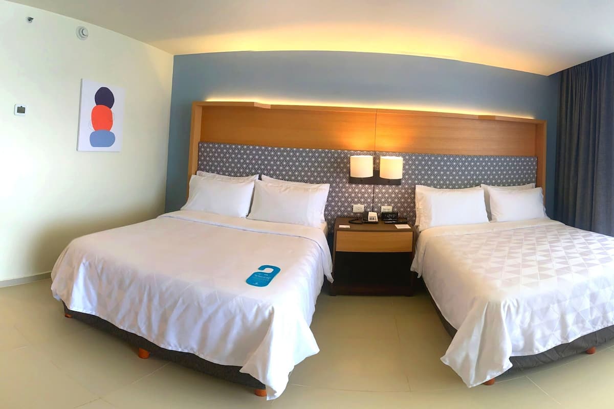 Habitación de hotel con dos camas, cabecero con diseño geométrico, mesita de noche con un reloj y aplique de pared.
