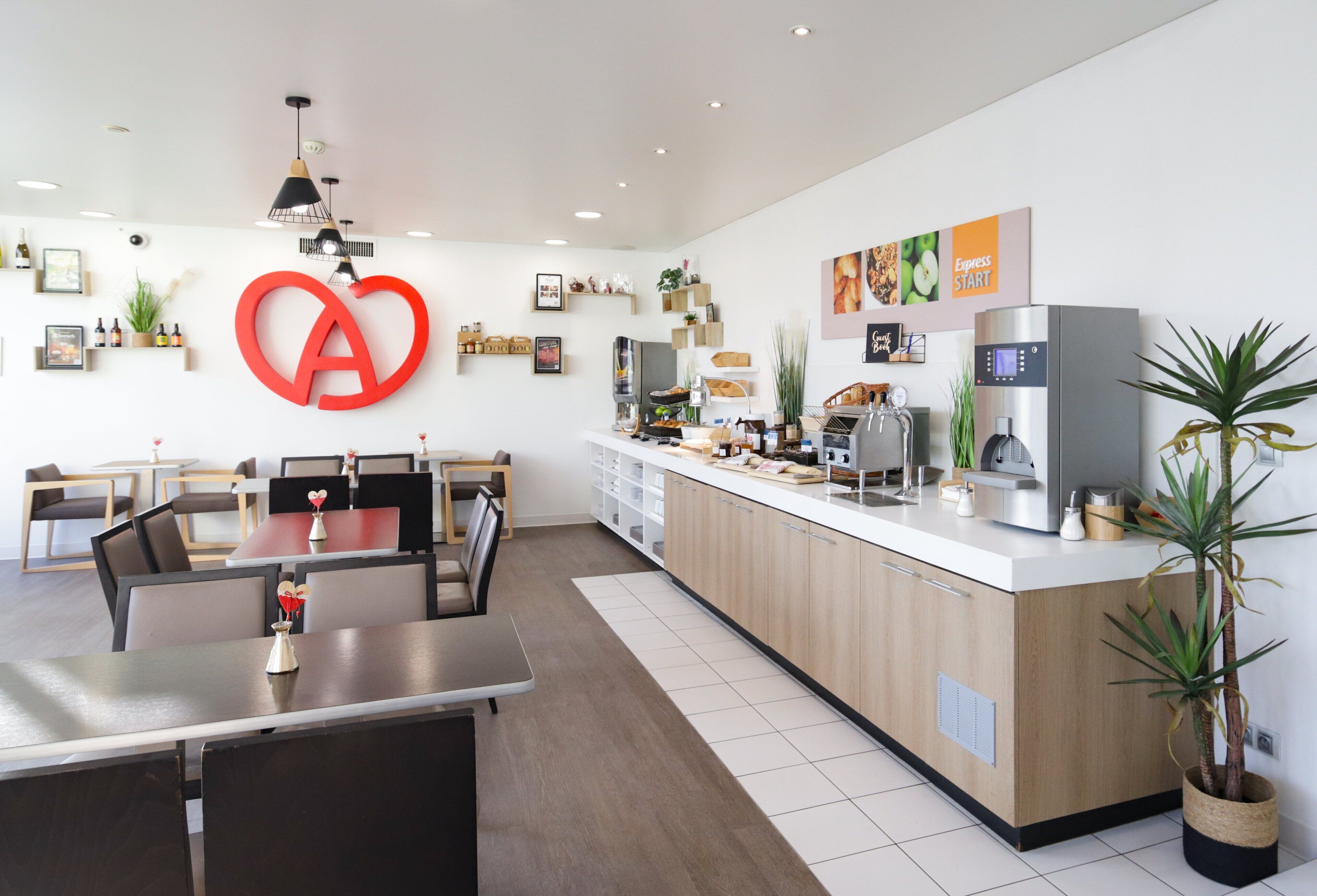 A modern hotel lobby with a central reception desk, a large heart-shaped logo on the wall, and a dining area with tables and chairs.
