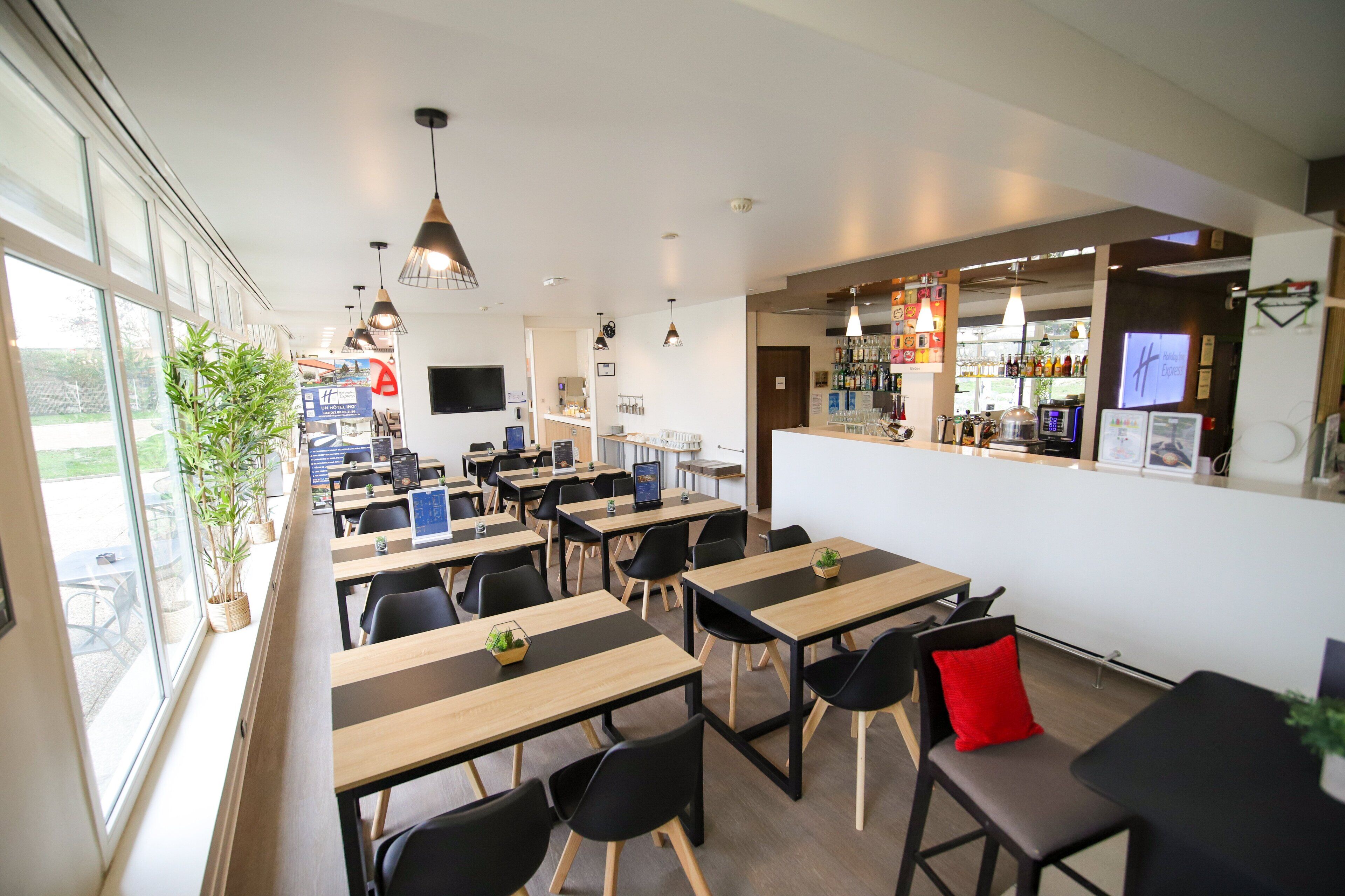 A modern restaurant with wooden tables and black chairs, a bar area with a TV, and potted plants.