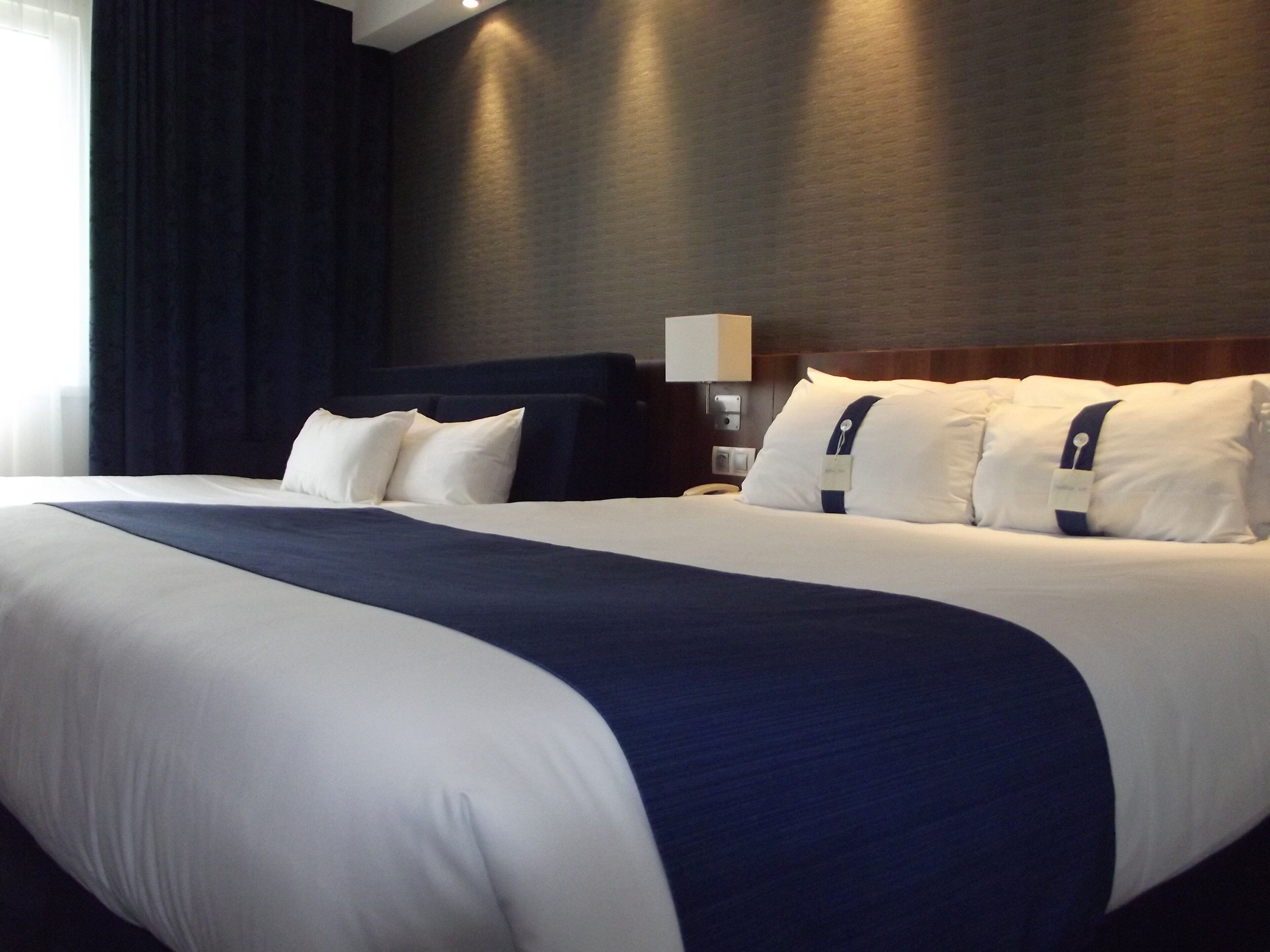 Una camera d'albergo con un letto grande, una testiera scura e un comodino con una lampada.