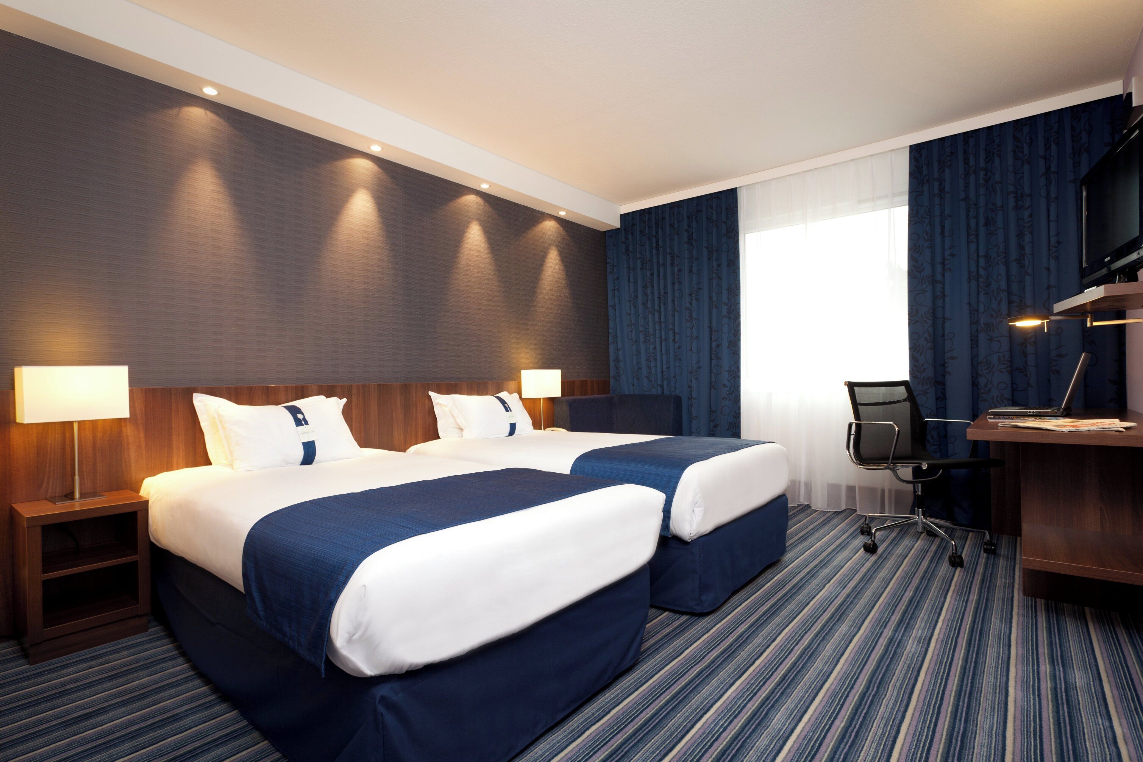 A hotel room with two beds, a desk with a chair, a TV, and a window with curtains.
