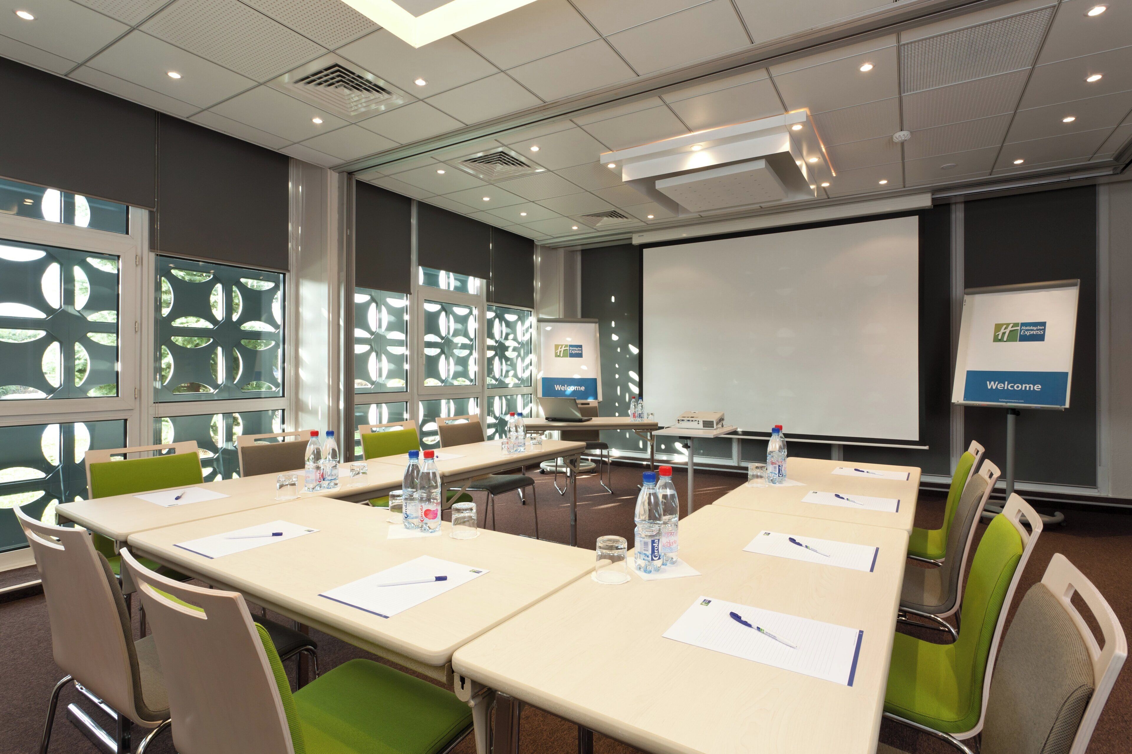 Una sala de conferencias con una pantalla grande, mesas con sillas, botellas de agua y un cartel que dice 'Welcome'.