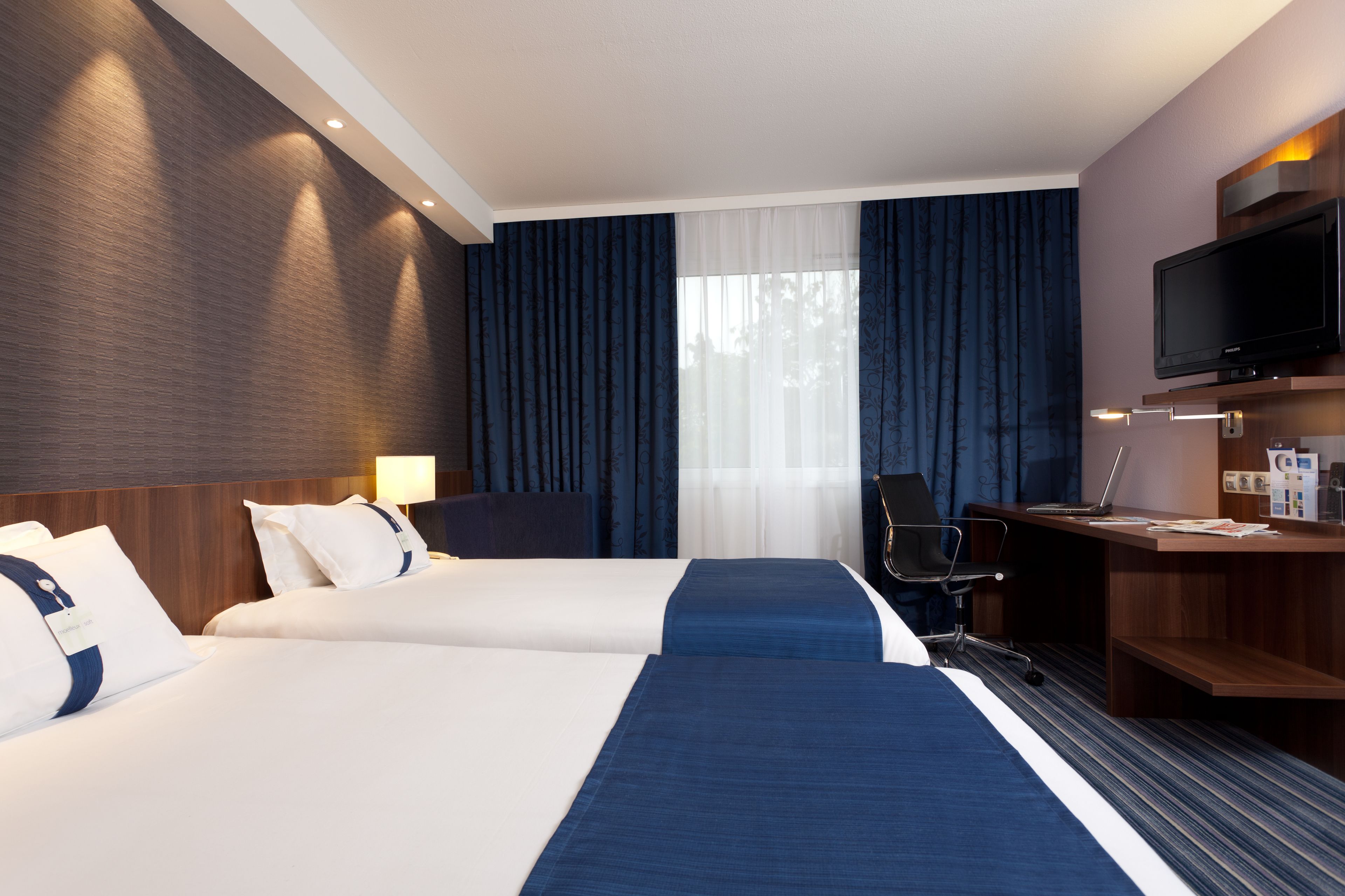 A hotel room with two beds, a desk, a chair, a TV, and a window with curtains.