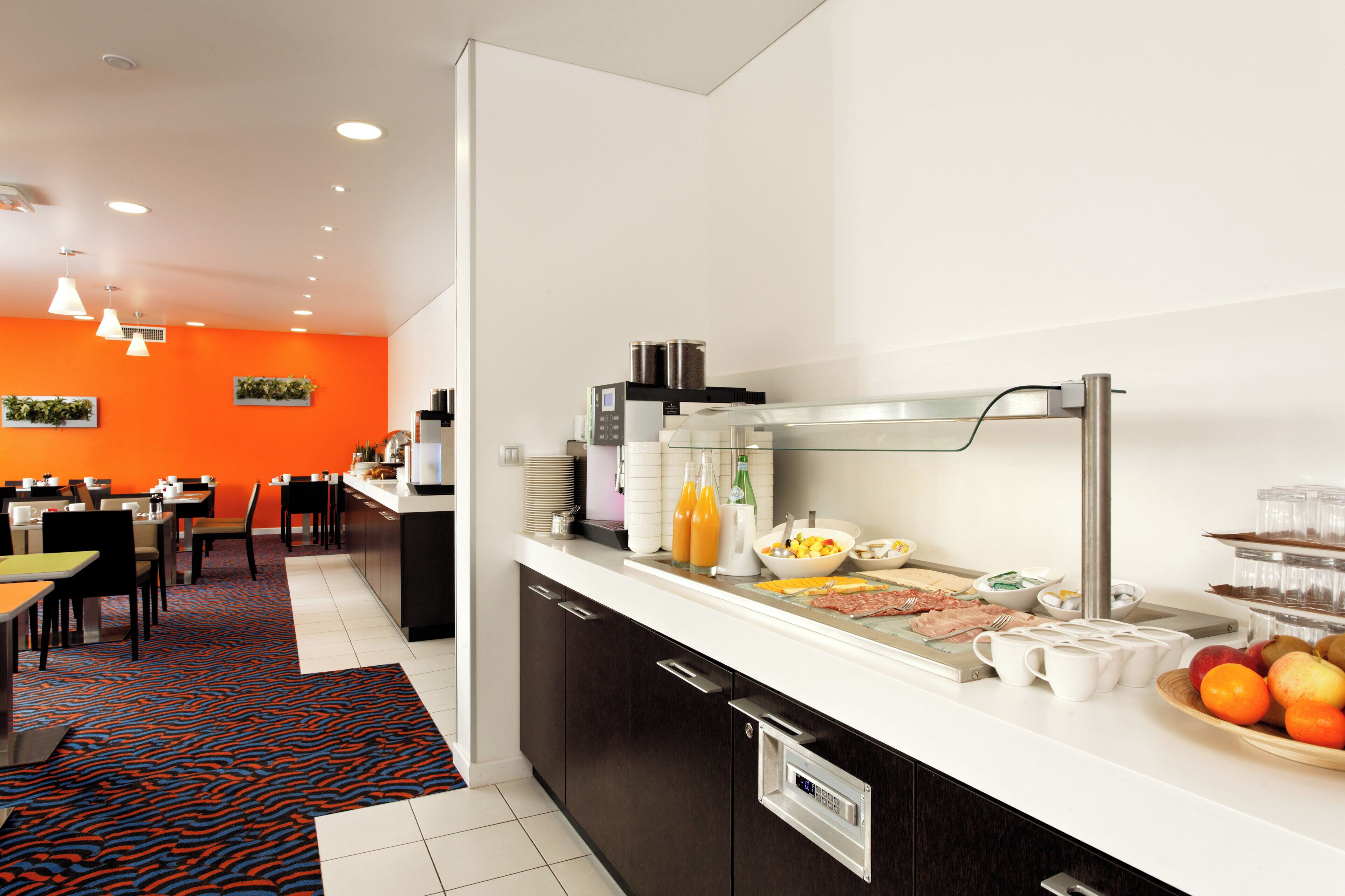 A modern hotel room with a breakfast bar, dining area, and a kitchen counter with various food items and beverages.
