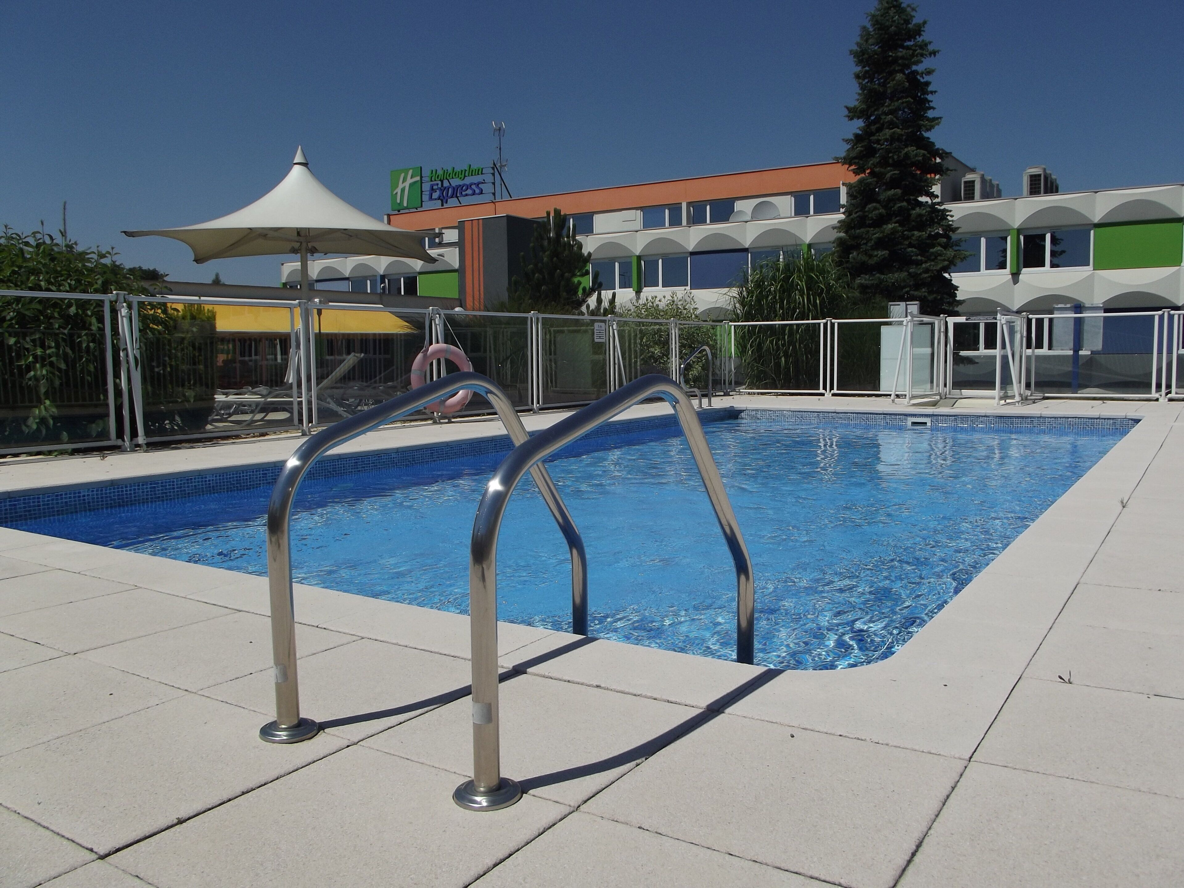 Une piscine avec des échelles en métal, un espace de repos couvert et, au fond, le bâtiment d’un hôtel Holiday Inn Express.